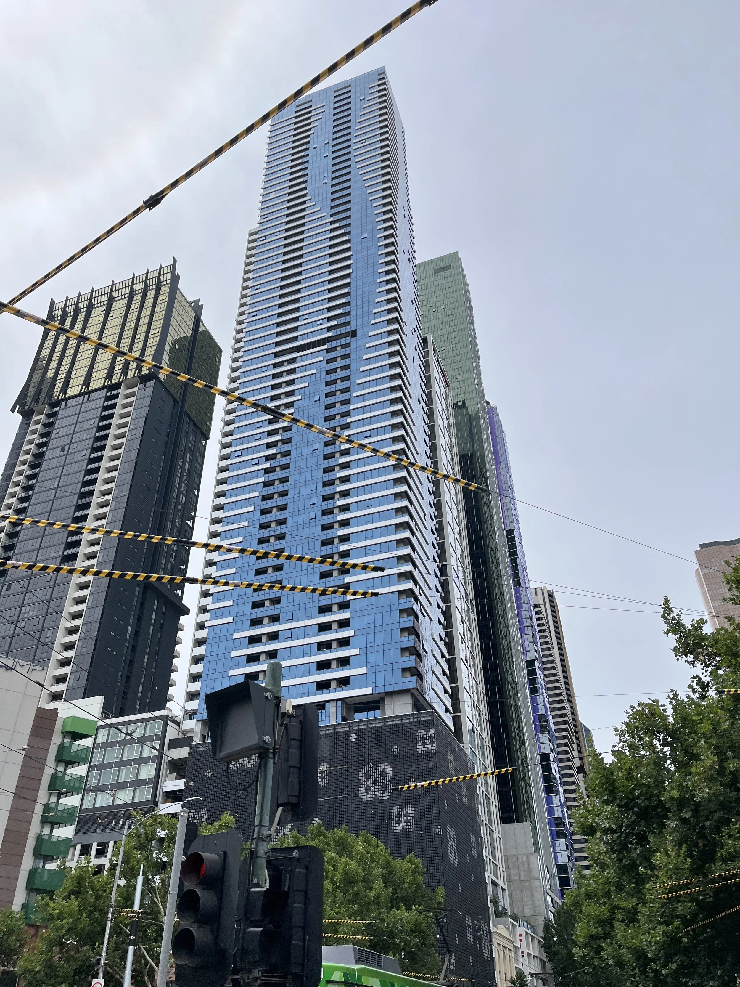 Tall blue and black skyscraper under a cloudy sky in a city, with traffic lights and trees in the foreground.