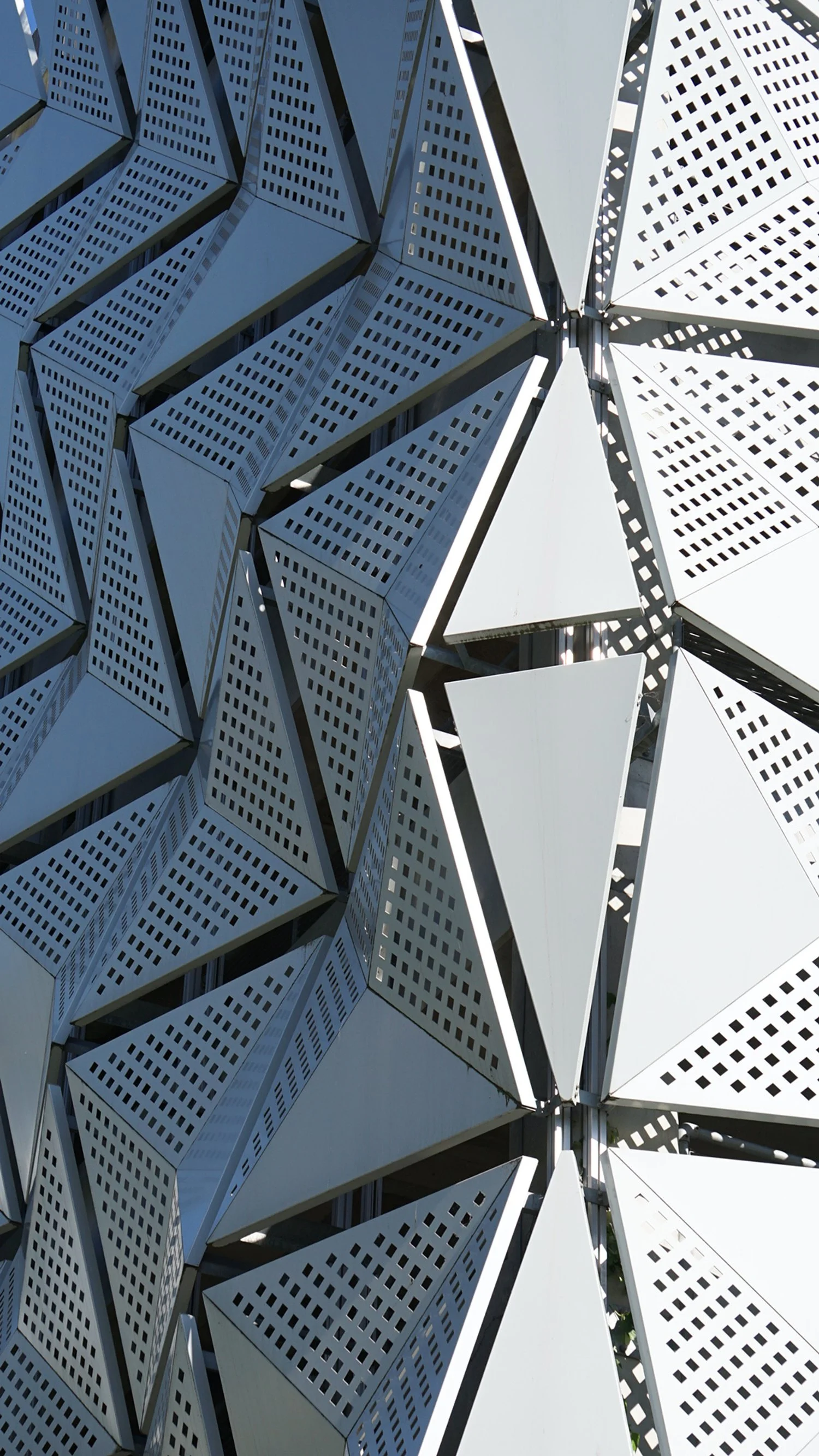 Close-up of a modern architectural facade composed of geometric, pyramid-shaped metallic panels with perforated surfaces, arranged in a repeating pattern.