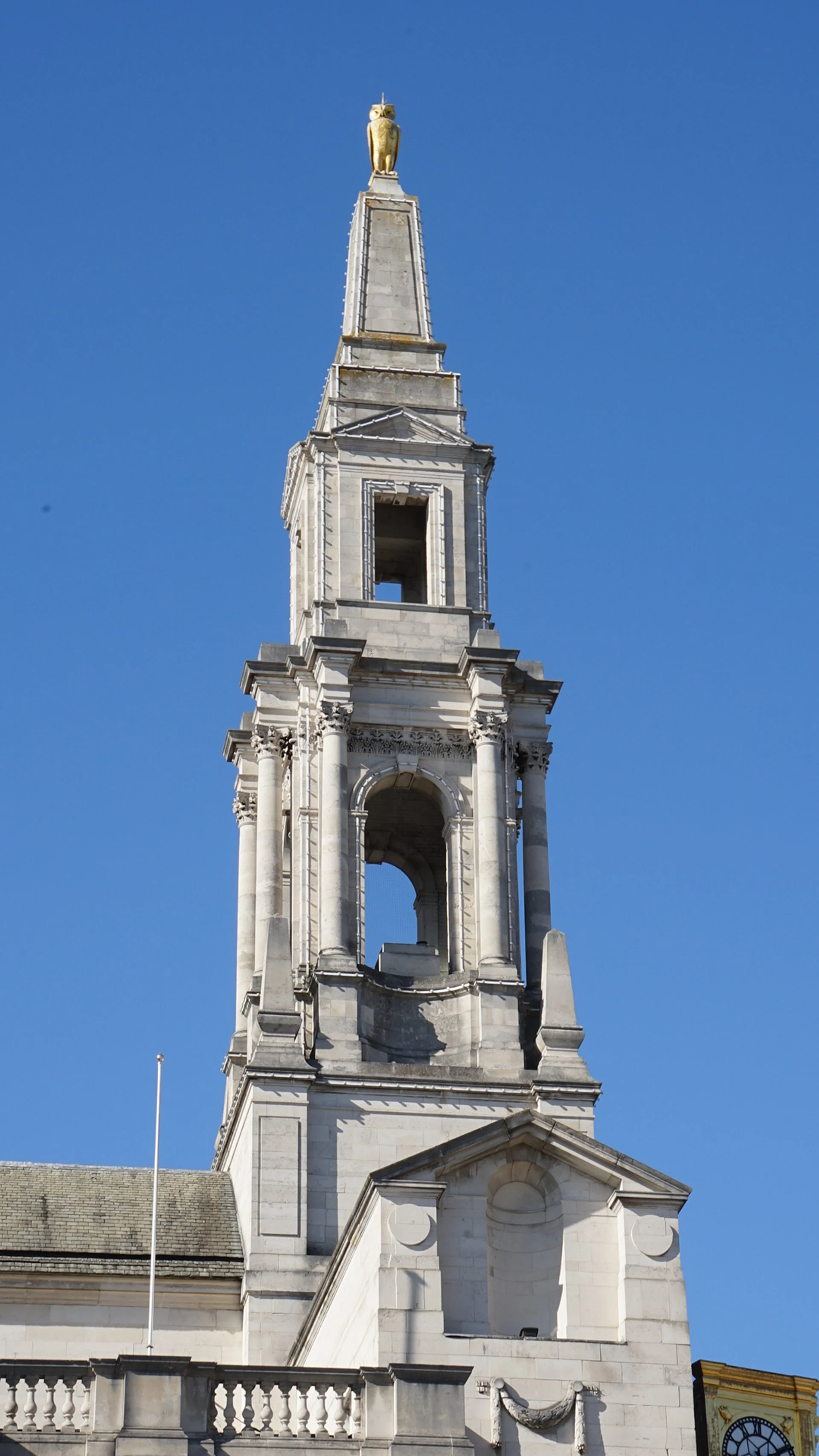Tall white stone church steeple with a golden owl on top, against a clear blue sky.