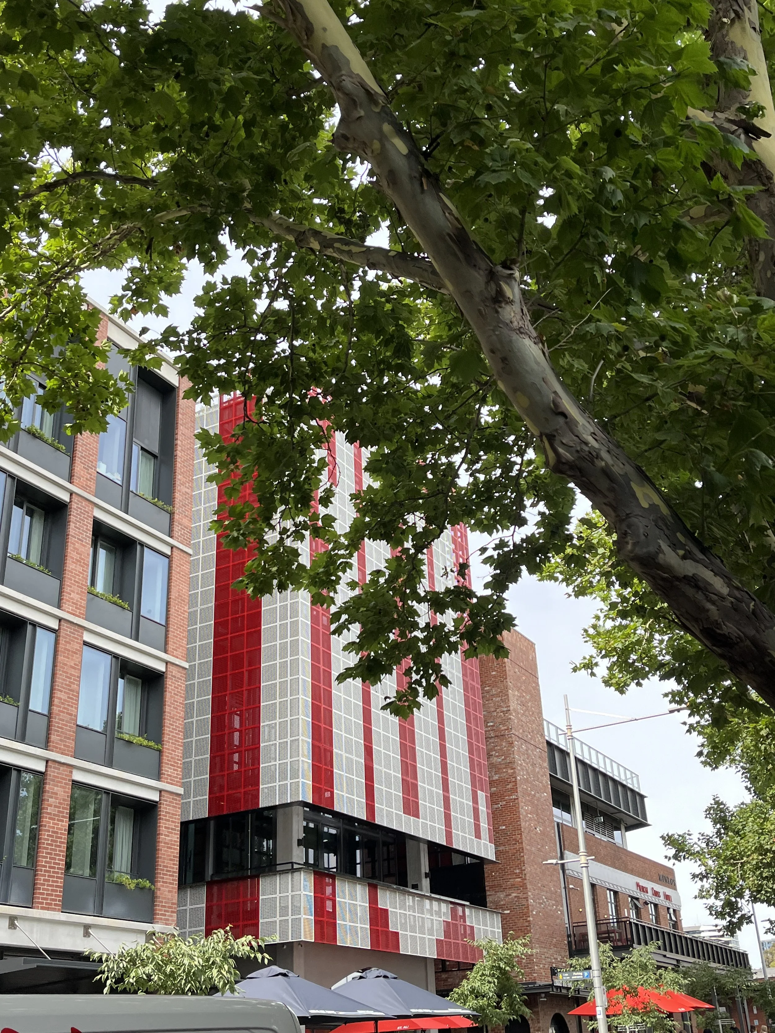 A view of a modern multistory building with a red and gray patterned facade, partially obscured by green leafy trees in an urban setting, with outdoor umbrellas and seating visible at street level.