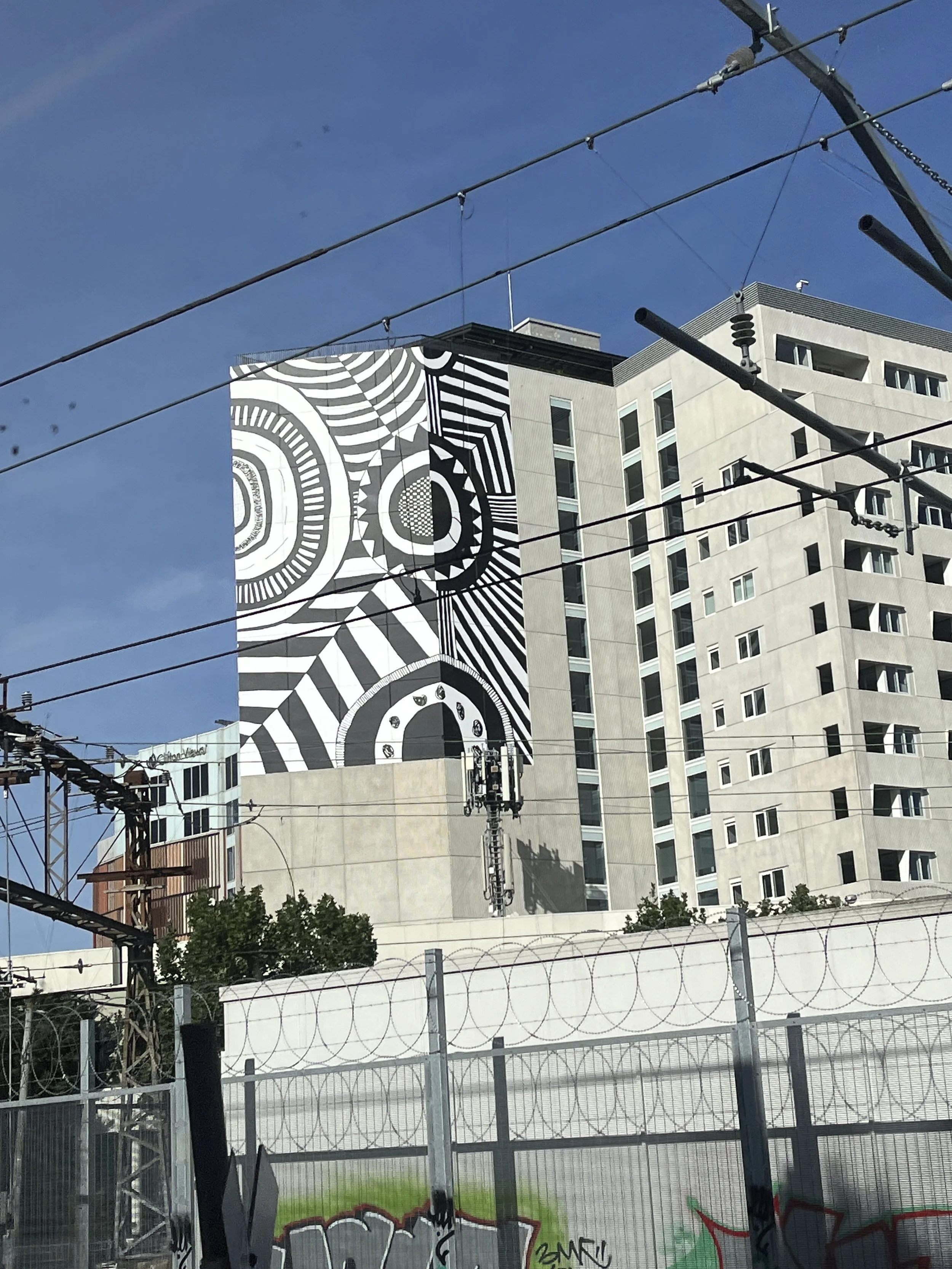 A tall building with a mural featuring black and white geometric patterns, set against a clear blue sky with power lines and a fence in the foreground.