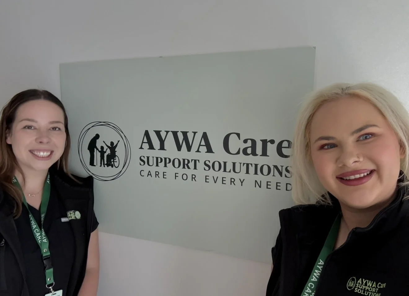 Two women smiling for a photo in front of an AYWA Care Support Solutions sign. Both are wearing black vests with green lanyards.