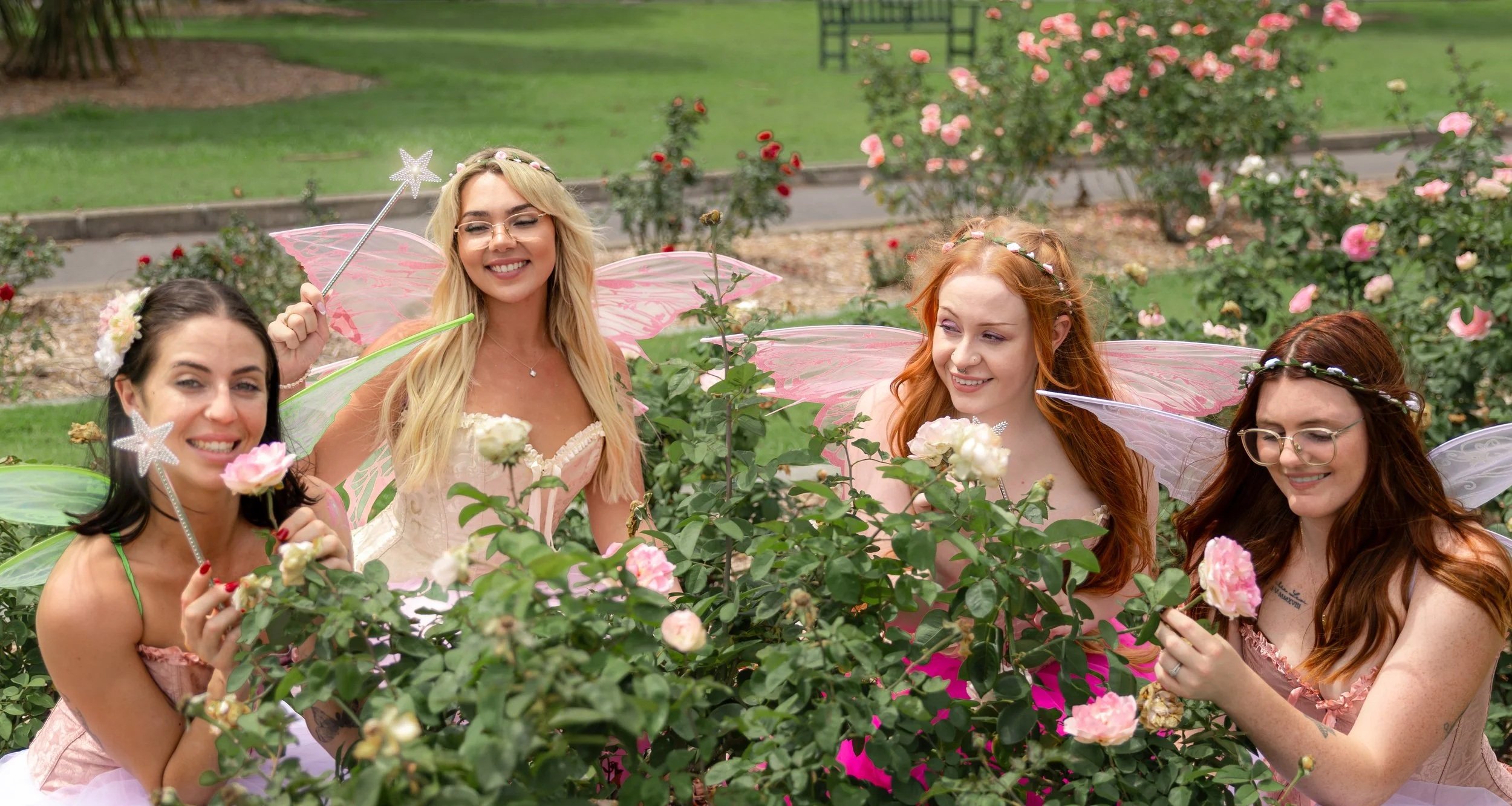 Four women dressed as fairies with pink and purple wings and fairy wands, smiling and enjoying a garden filled with pink roses.