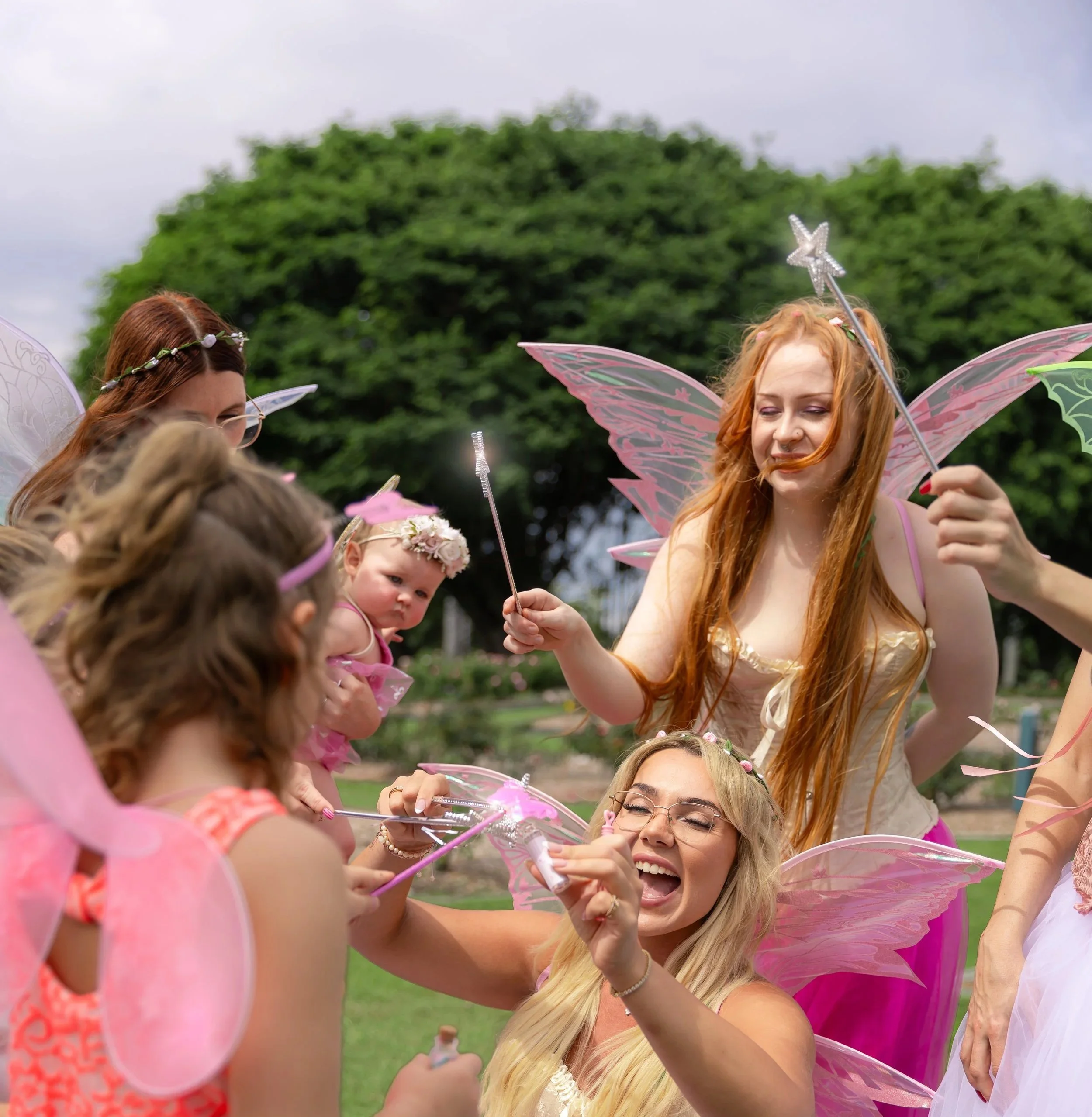 Group of children and women dressed as fairies with wings and wands outdoors, engaging in a fairy-themed activity.