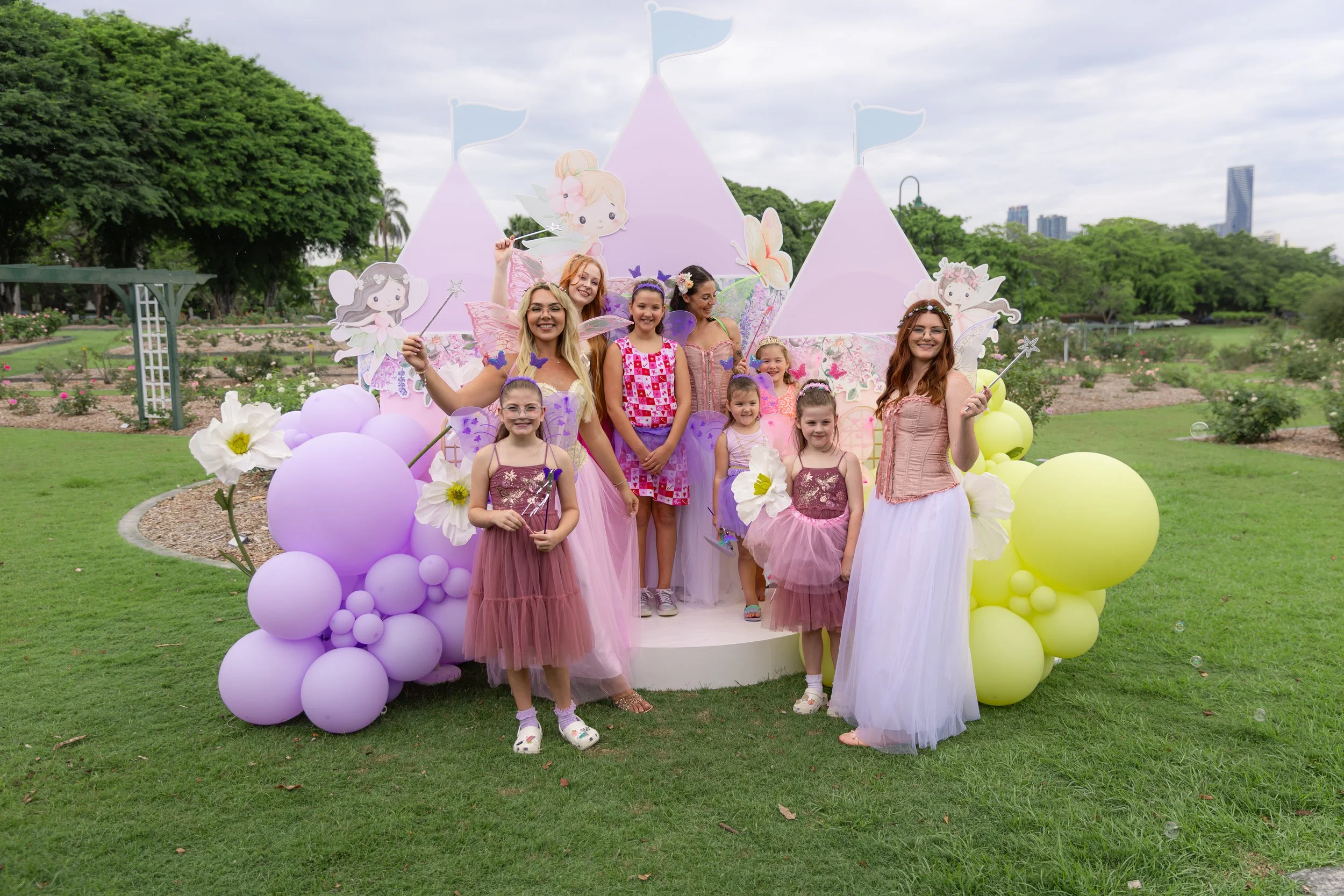 Young girls and adult women celebrate a fairy-themed birthday party outdoors with pastel-colored decorations, balloons, and fairy wings in a park with trees and city buildings in the background.