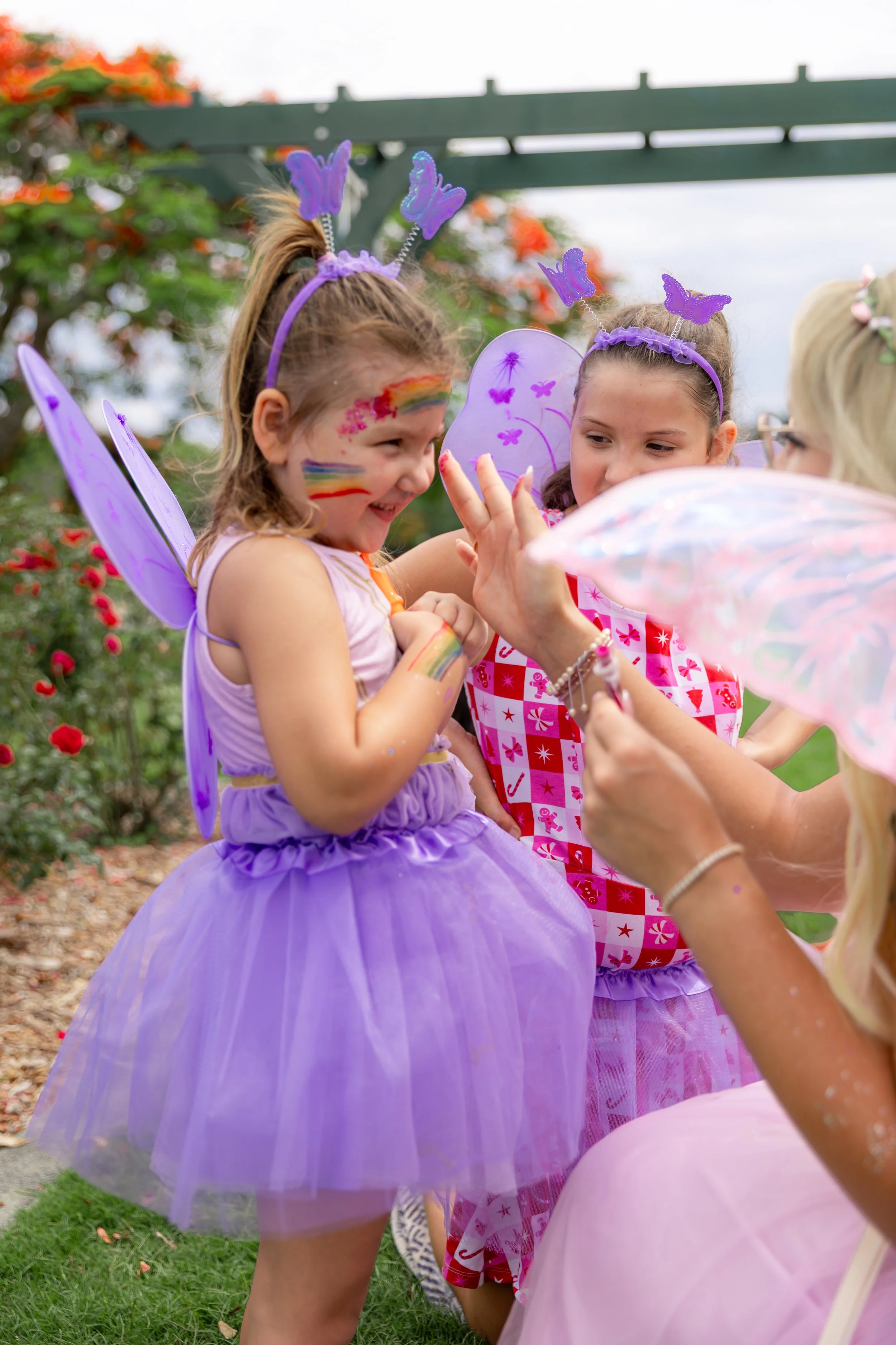 Young girls dressed as fairies, wearing purple tutus and fairy wings, enjoying a playful moment outdoors during a festive event.