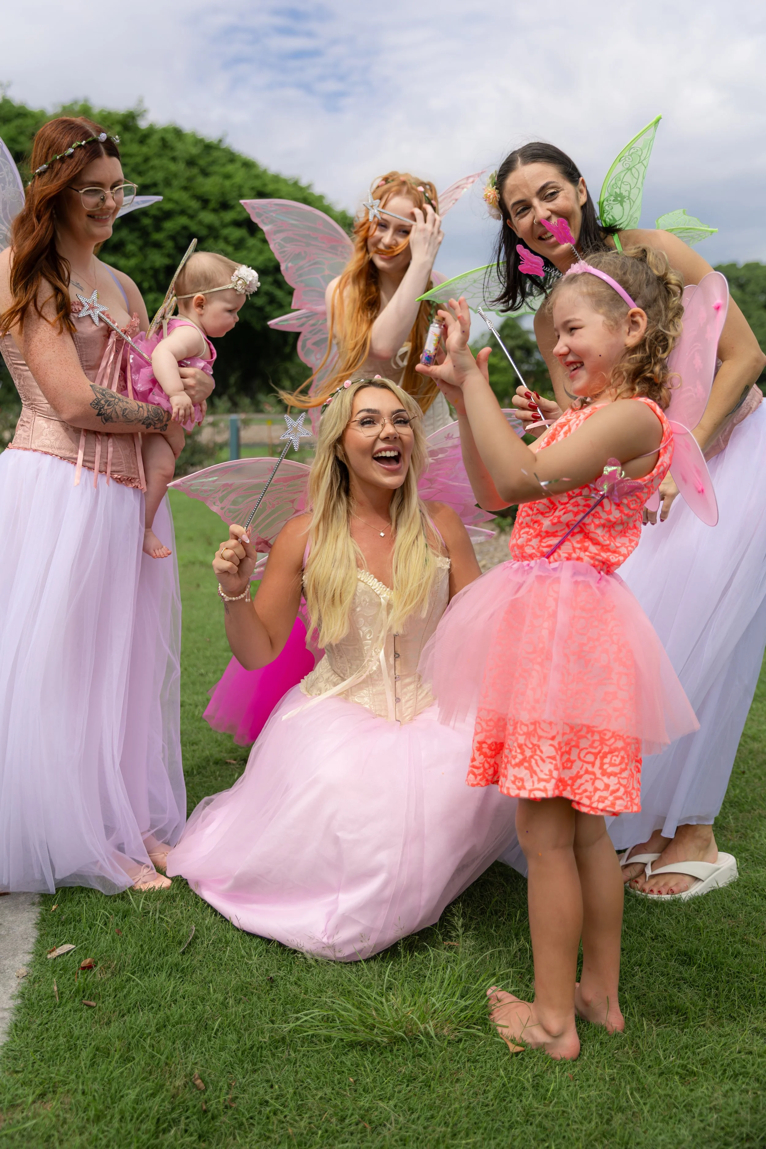 Group of women and children dressed as fairies with wings and wands, celebrating outdoors on a grassy field.