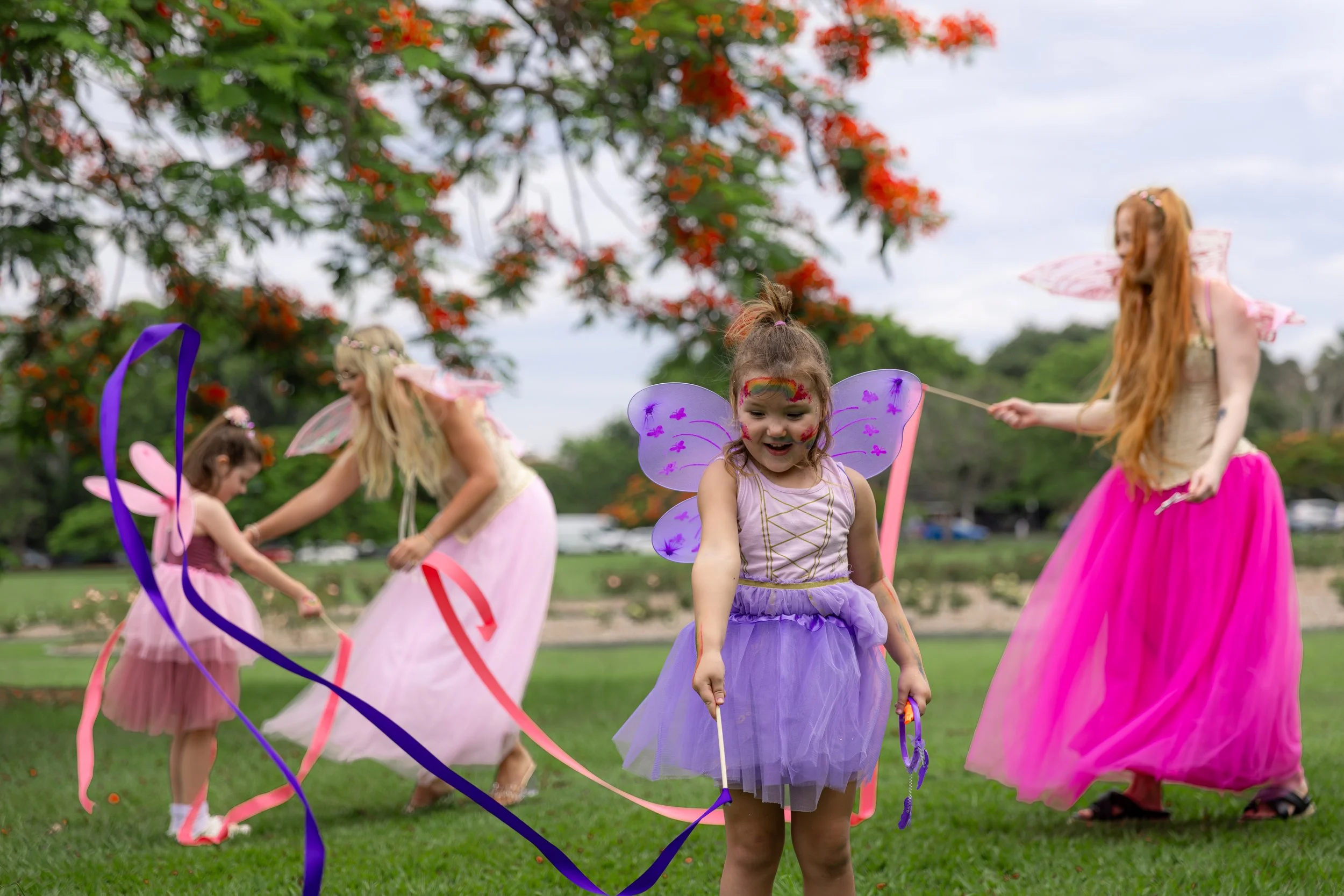 Young girls dressed as fairies with wings and fairy costumes playing and having fun outdoors on green grass during daytime.