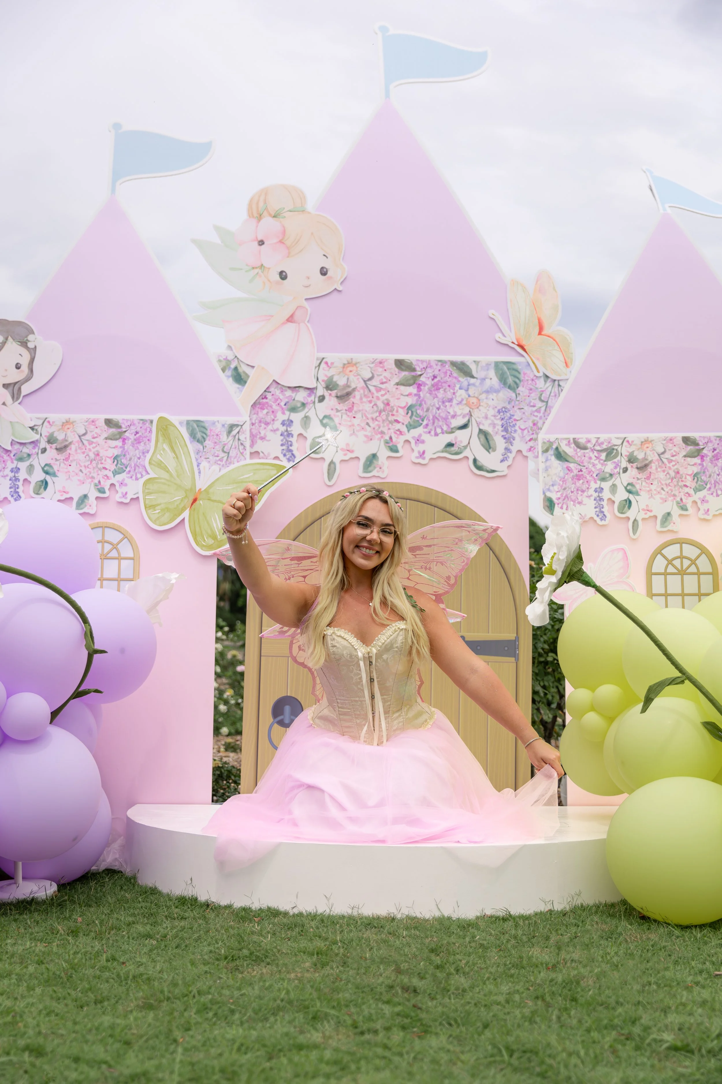Woman in a pink fairy costume sitting on a decorated outdoor fairy tale-themed set with balloons, floral decorations, and cartoon fairy illustrations.