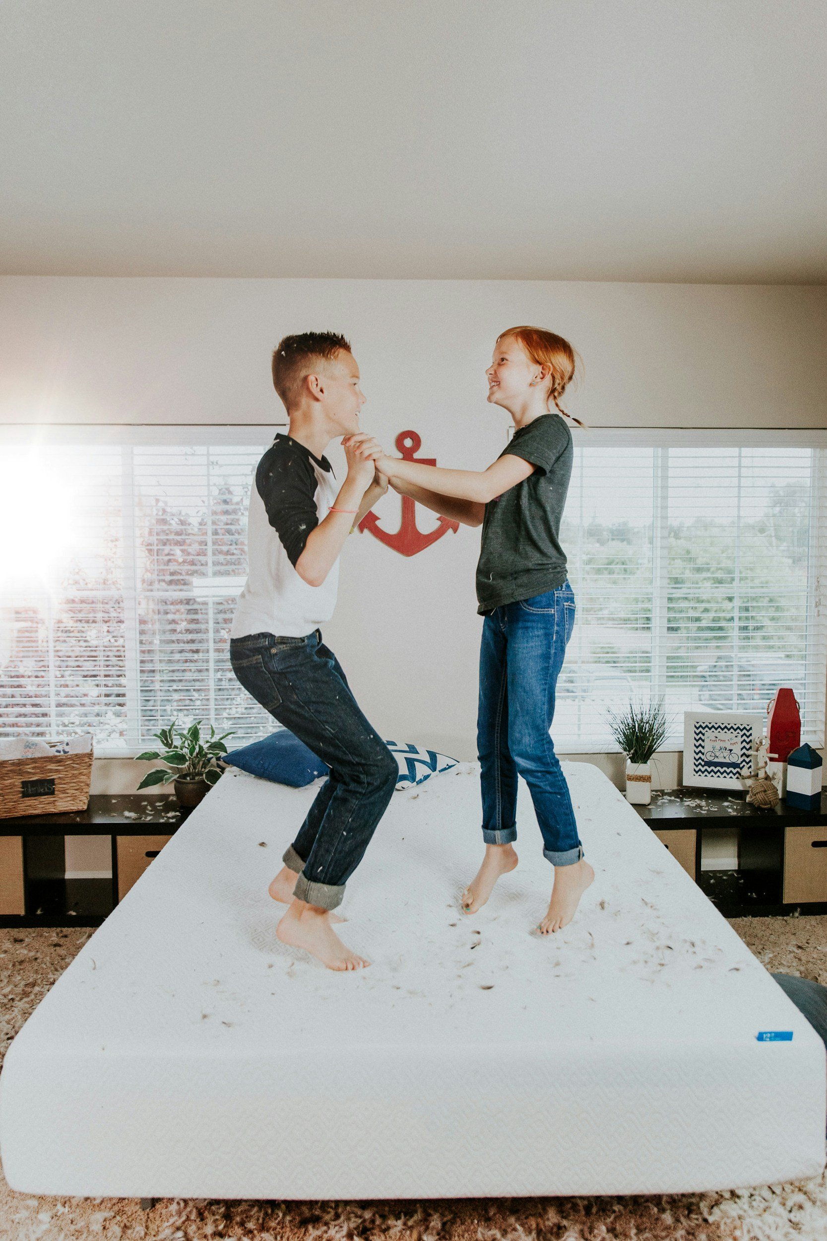 Two children, a boy and a girl, playing and jumping on a messy bed indoors, with plants and decorations in the background.