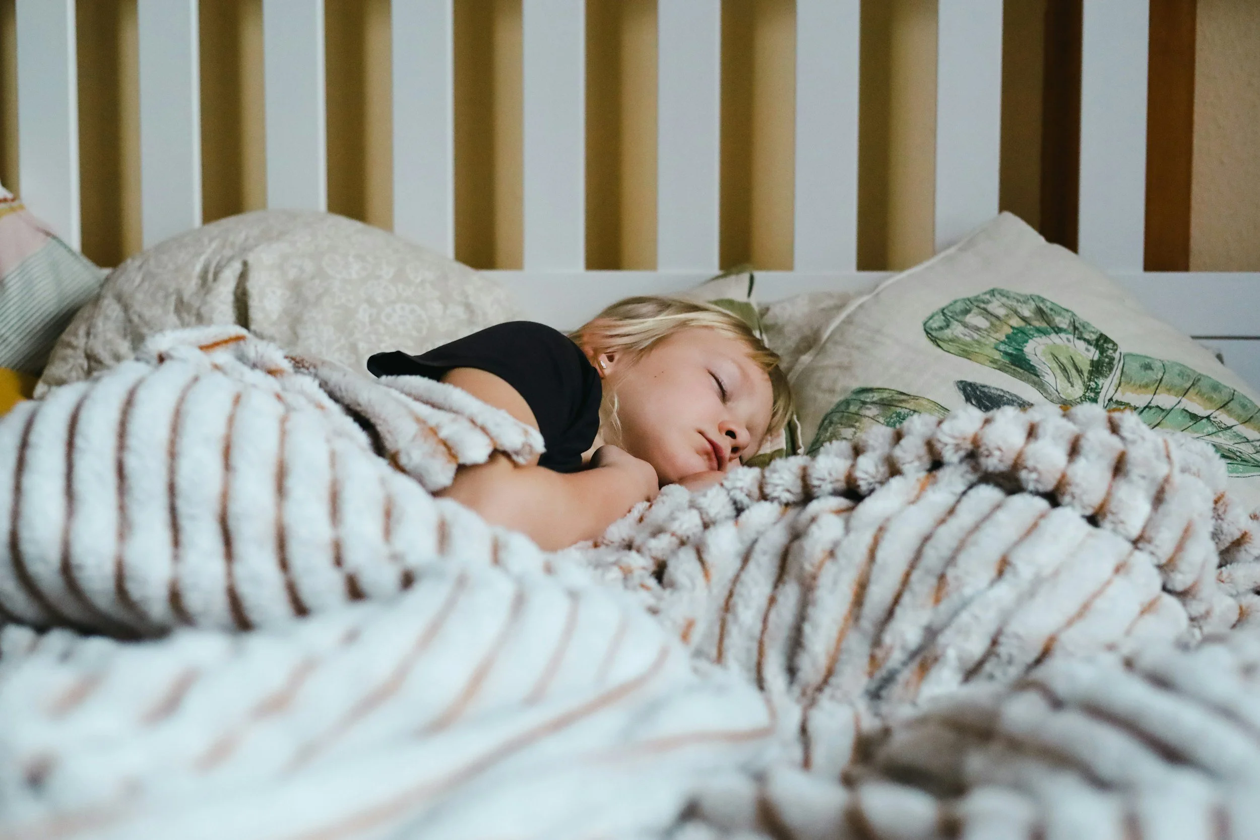 A young girl with blonde hair sleeping peacefully in bed, covered with a striped blanket, with a pillow featuring a leaf design behind her.