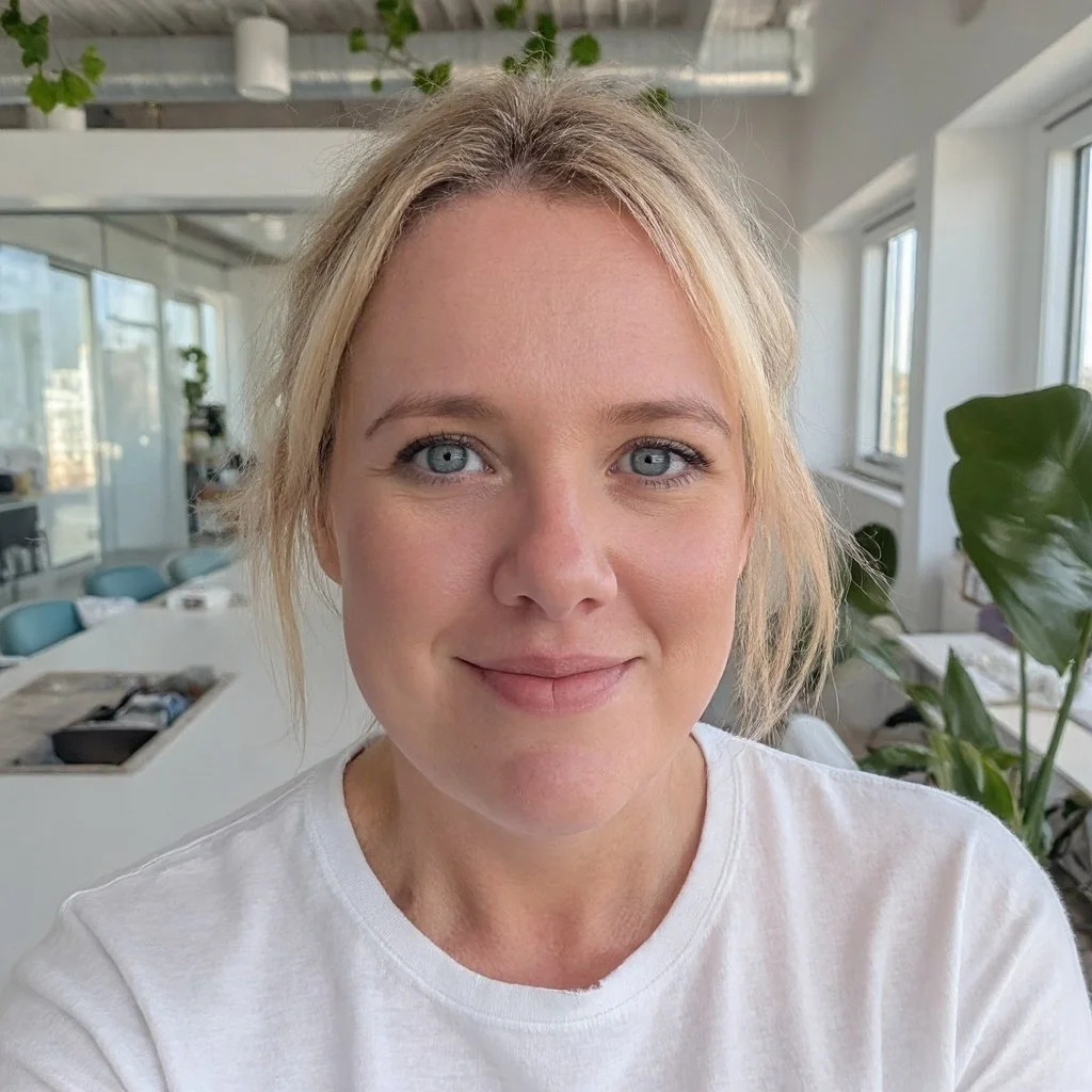 Close-up of a young woman with blonde hair and blue eyes smiling in an office environment with plants and large windows.