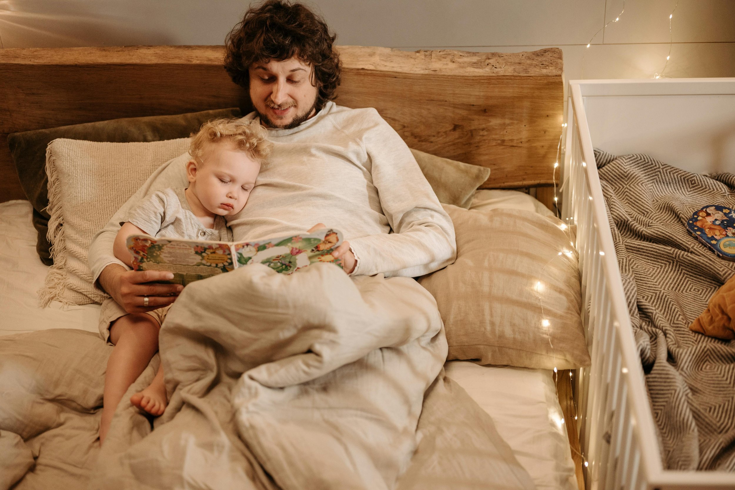 A man and young boy are lying in bed, reading a colorful book together. The boy appears sleepy, resting against the man, who is smiling. The bed has beige sheets and pillows, with fairy lights on the headboard and a fairy-tale themed puzzle on a nearby bed or dresser.