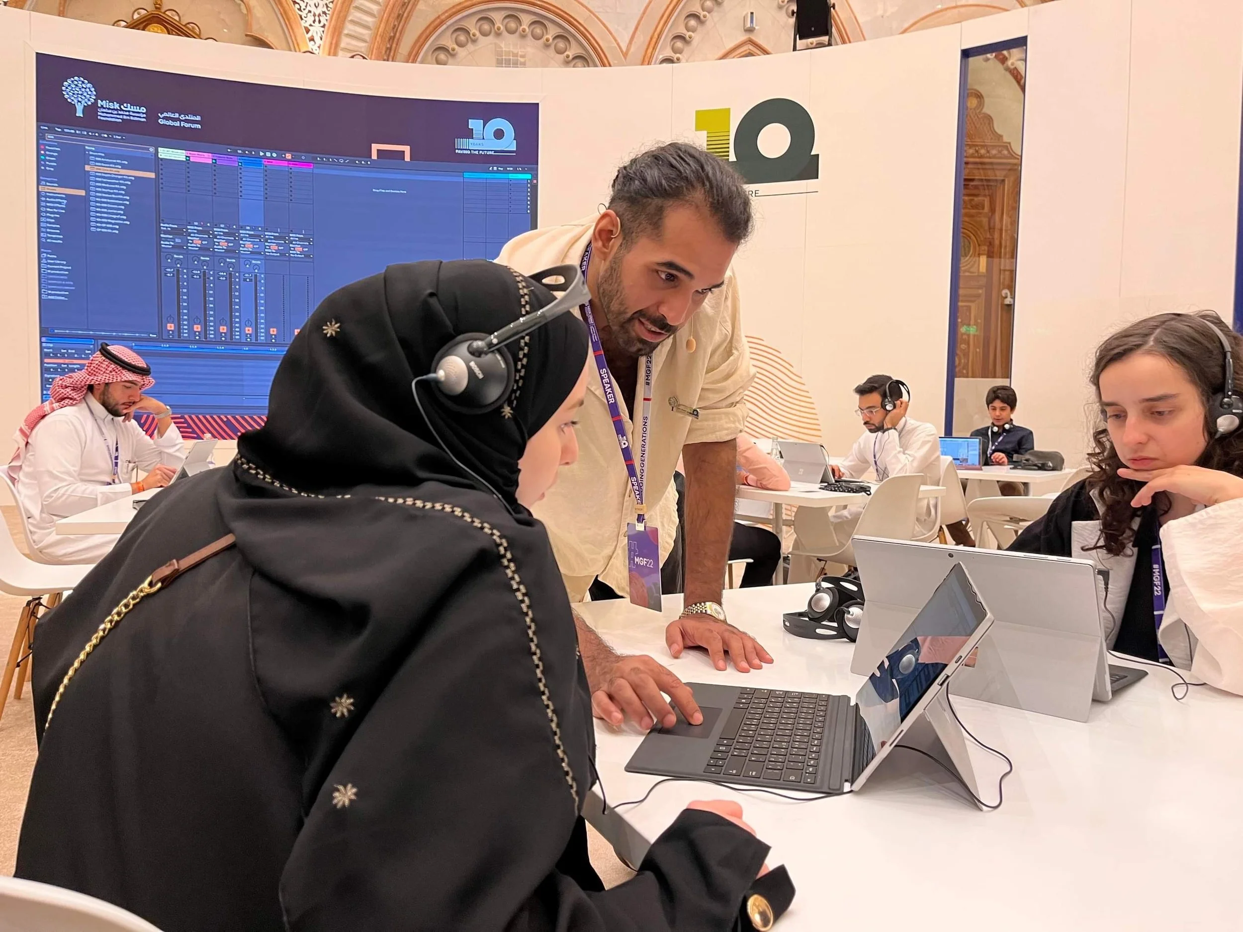 People working at computers in a conference or workshop setting, with a large display screen in the background showing digital audio workstation software, and a large decorative ceiling in the venue.