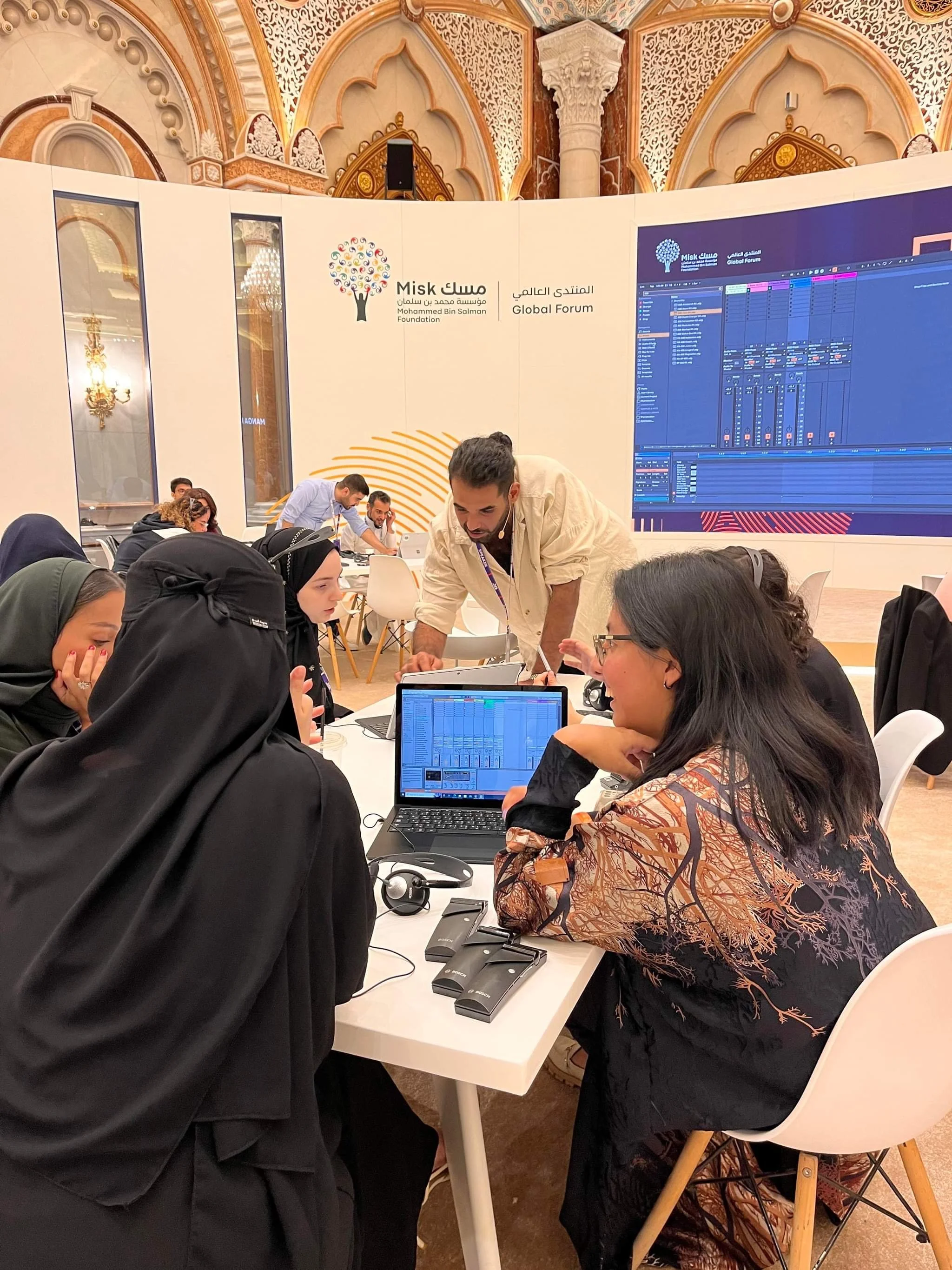 People attending a conference or workshop at a large, ornate hall, with a large screen displaying music software. A man is assisting women seated at a white table with laptops and headphones.