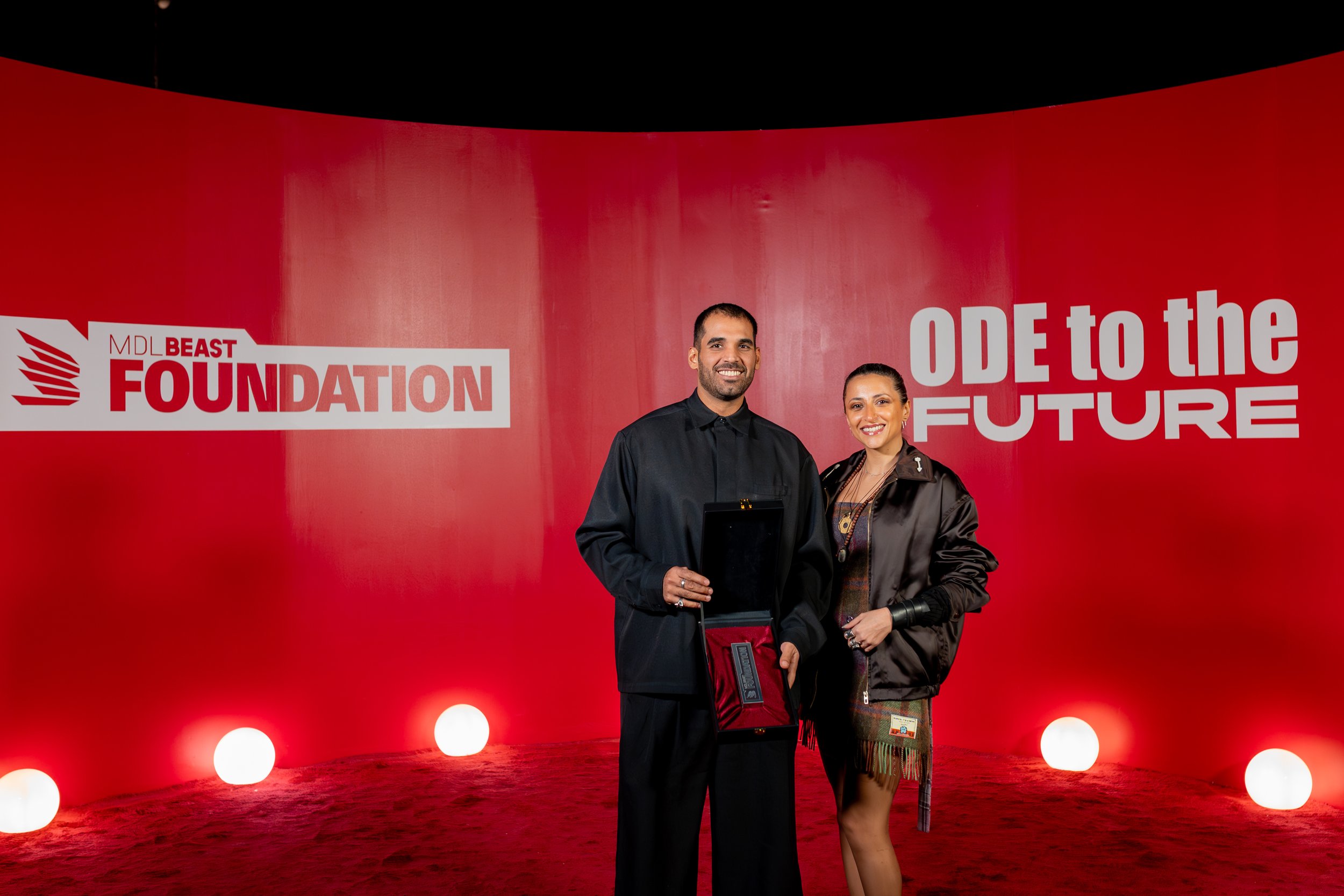 Two people standing in front of a red backdrop with the text 'MDL Beast Foundation' and 'Od To The Future'. They are smiling, with one person holding an award or plaque.