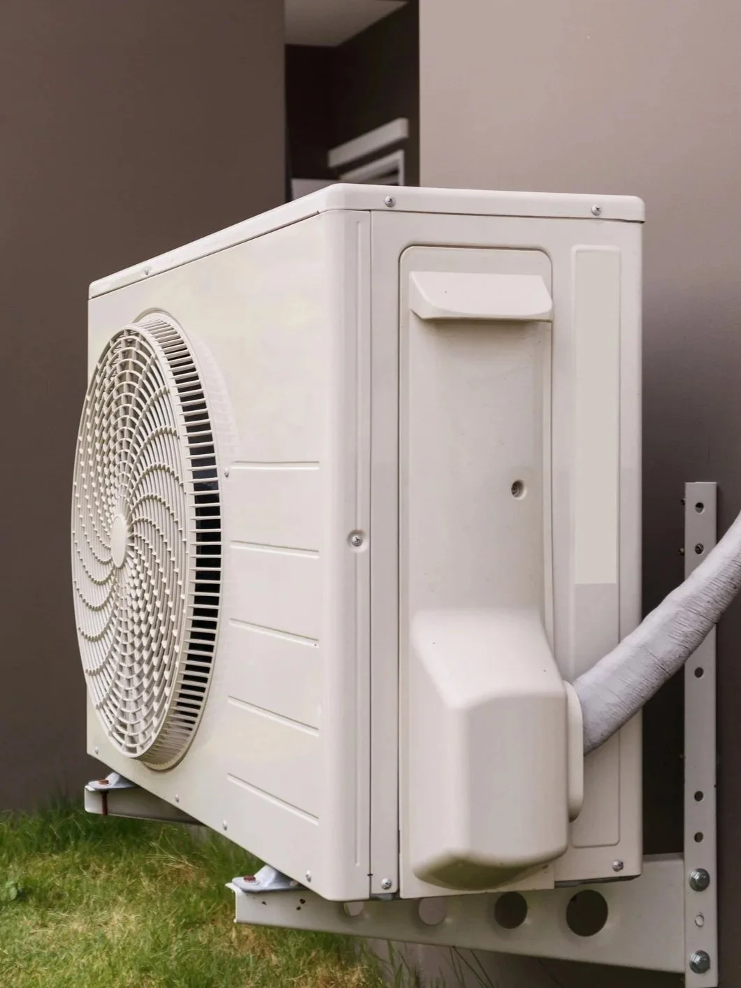An outdoor air conditioning unit mounted on a wall with a fan on the front and a white insulated pipe connected to the right side.