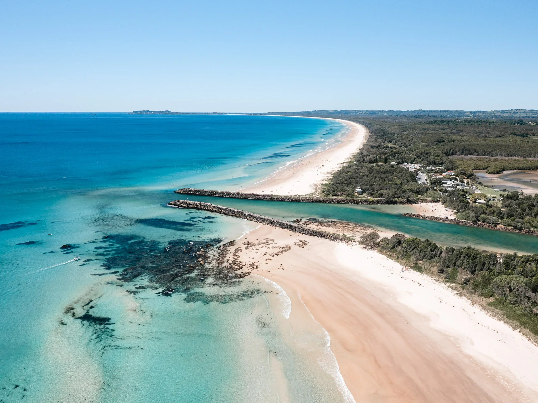 Aerial View of Brunswick Heads Breakwall, Brunswick Heads, Northern Rivers, Northern NSW. Aerial view of a sandy beach with calm blue ocean waves, a breakwater, and some rocks in shallow water, with greenery and trees along the shoreline.