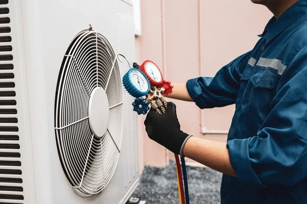 Technician wearing blue uniform and black gloves checking refrigerant gauges connected to an HVAC unit.