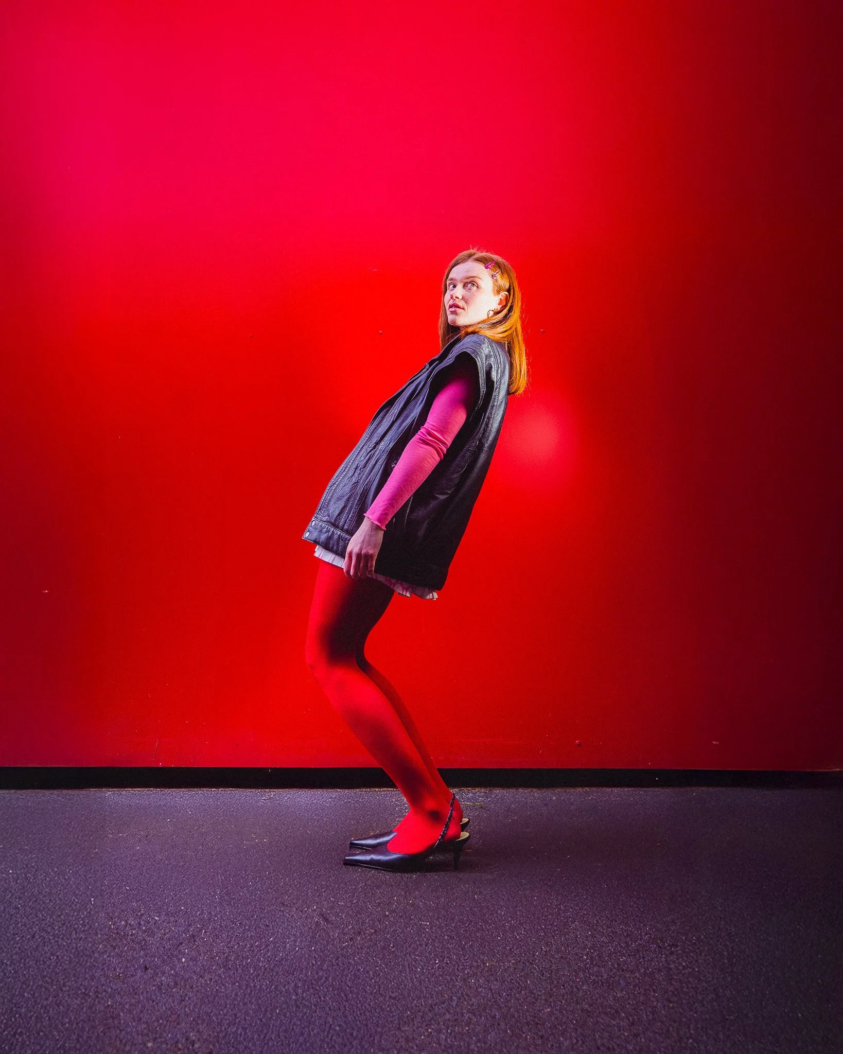 Femme debout contre un mur rouge, portant un manteau en cuir, une robe, des collants rouges et des talons hauts