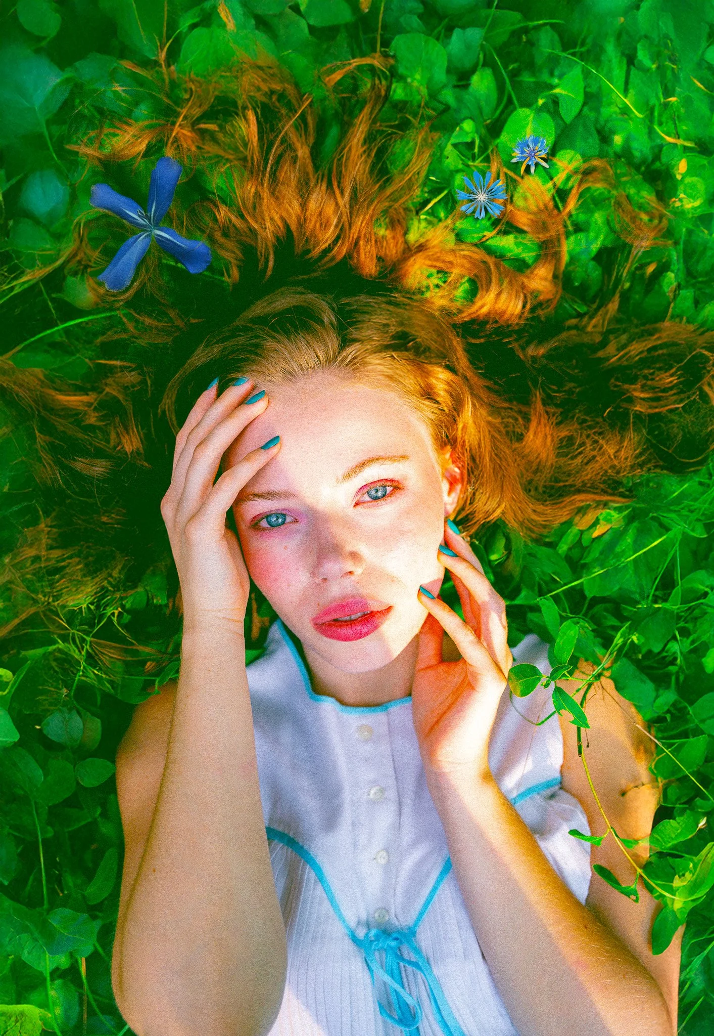 Jeune femme aux cheveux roux allongée dans un champ de verdure avec des fleurs bleues, regardant le haut avec son visage et ses mains près du visage.