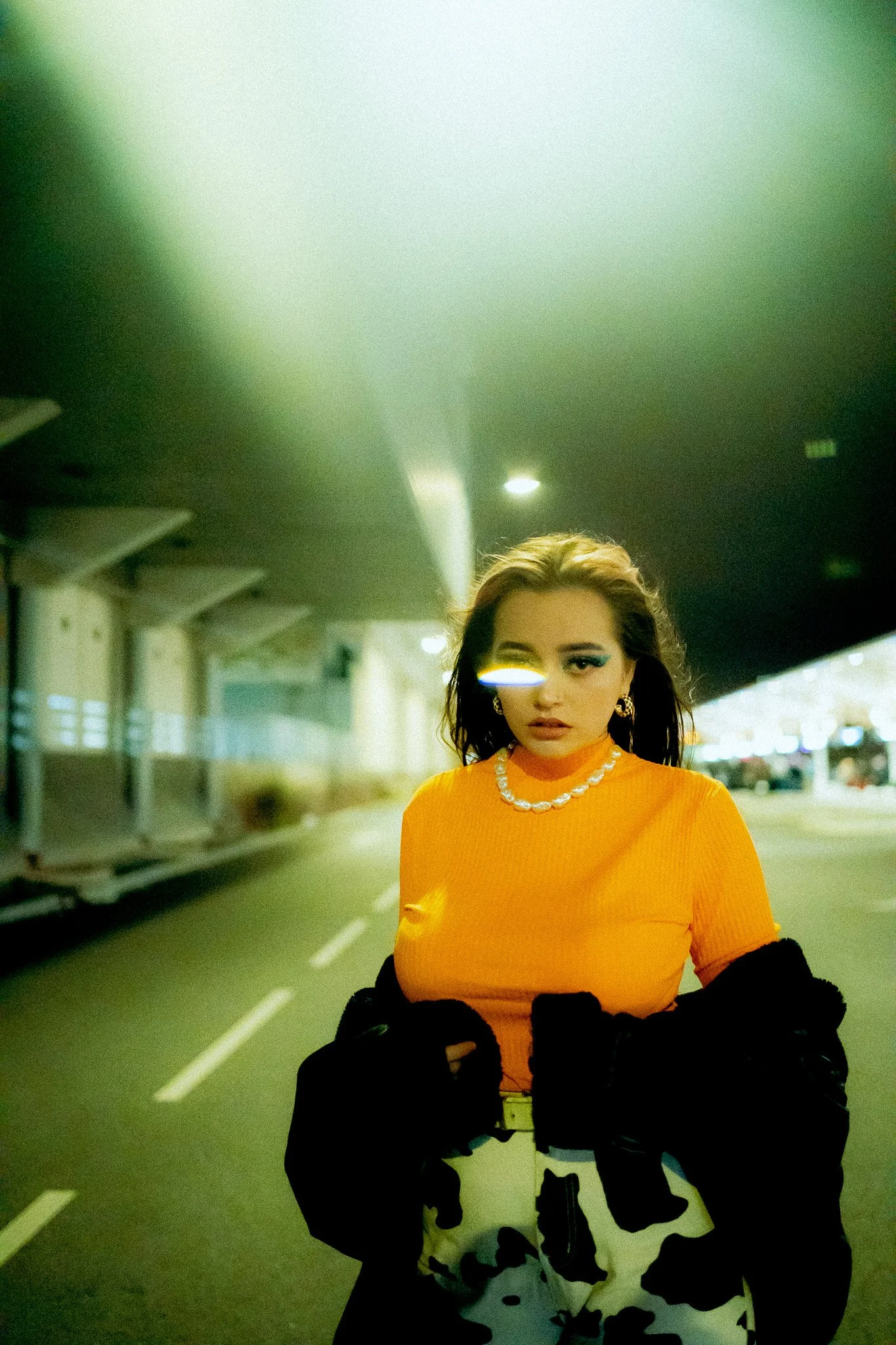 Une femme portant un haut orange, une veste noire, et un pantalon à motif noir et blanc, se tient dans une surface couverte sous un pont ou une structure architecturale similaire. Elle porte un collier et des boucles d’oreilles en or, avec des cheveu