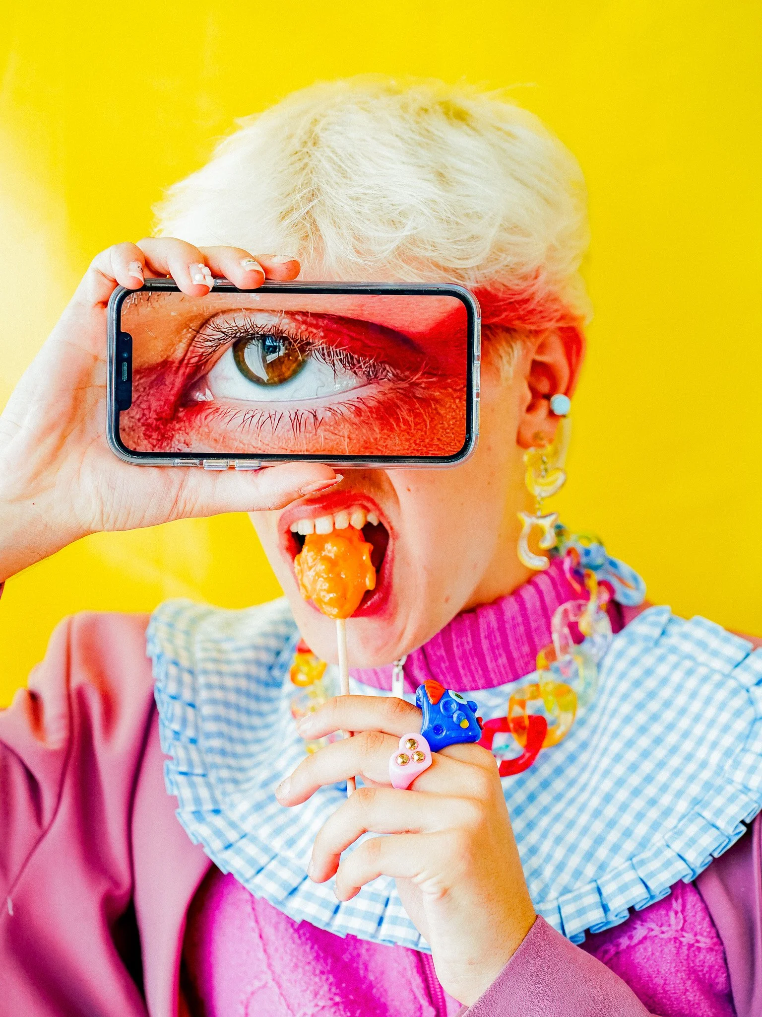Jeune personne blonde portant des bijoux colorés, mangeant une sucette, tenant un téléphone avec une image d'un œil en gros plan, vêtements colorés, fond jaune.