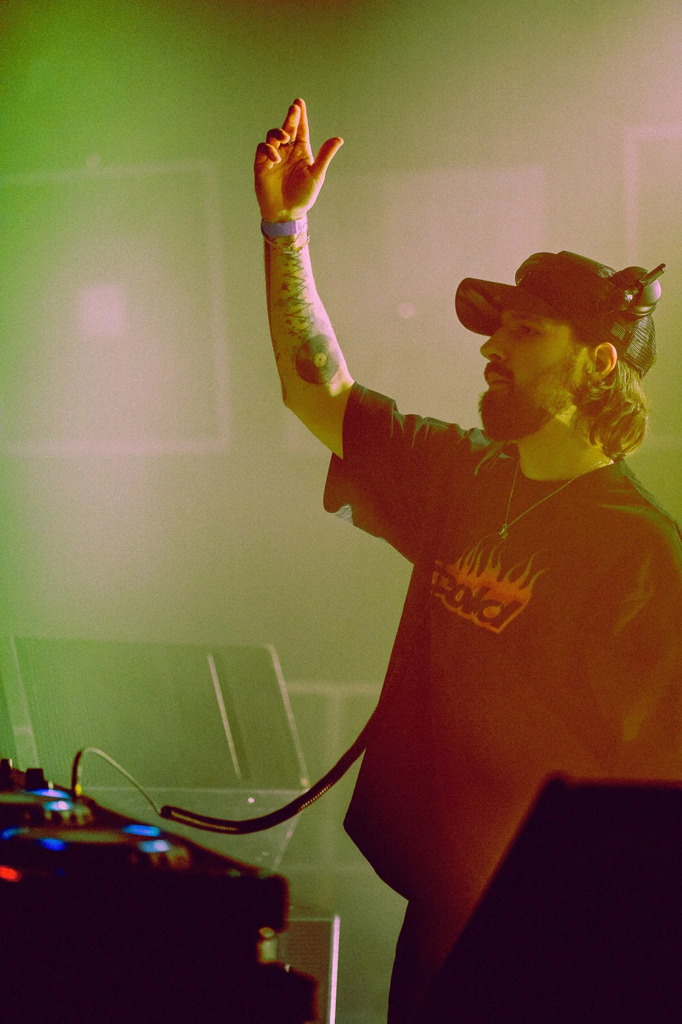 Un DJ avec un casque de réalité virtuelle sur la tête, portant un t-shirt noir avec un design de flammes, levant une main en l'air, se trouvant derrière un équipement de DJ, dans une ambiance de fête avec un éclairage coloré.