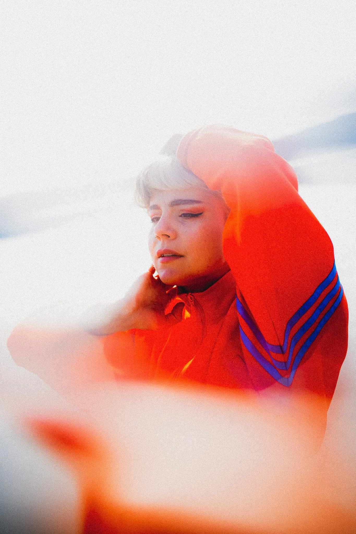 Une jeune femme avec des cheveux courts et blond platine porte une veste orange avec des bandes bleues et regarde pensivement, avec un ciel nuageux en arrière-plan.
