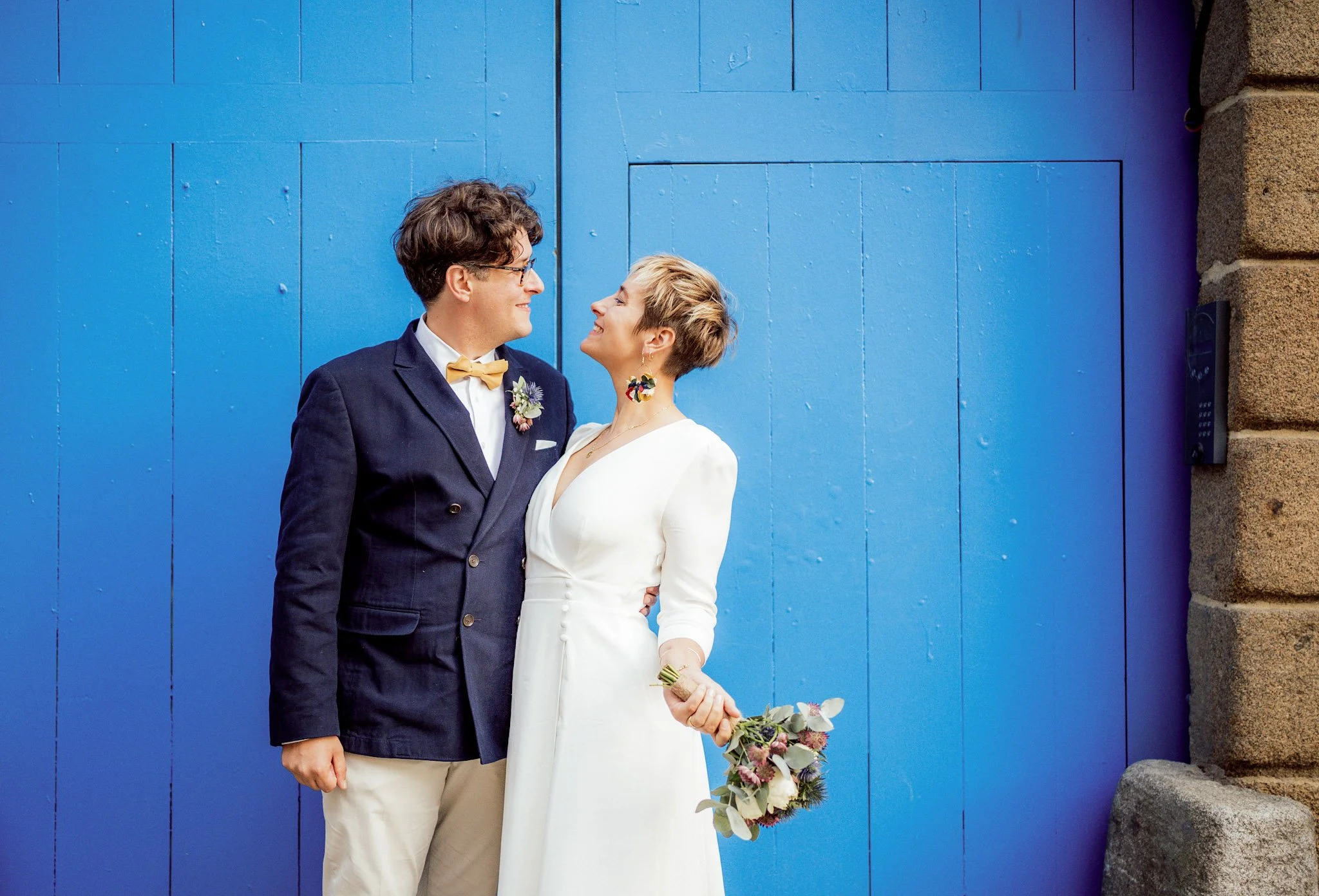 Un couple de mariage pose devant un mur bleu, la femme en robe blanche et le homme en costume foncé avec une chemise blanche, une cravate jaune et une boutonnière, tenant un bouquet de fleurs. Ils se regardent avec amour.