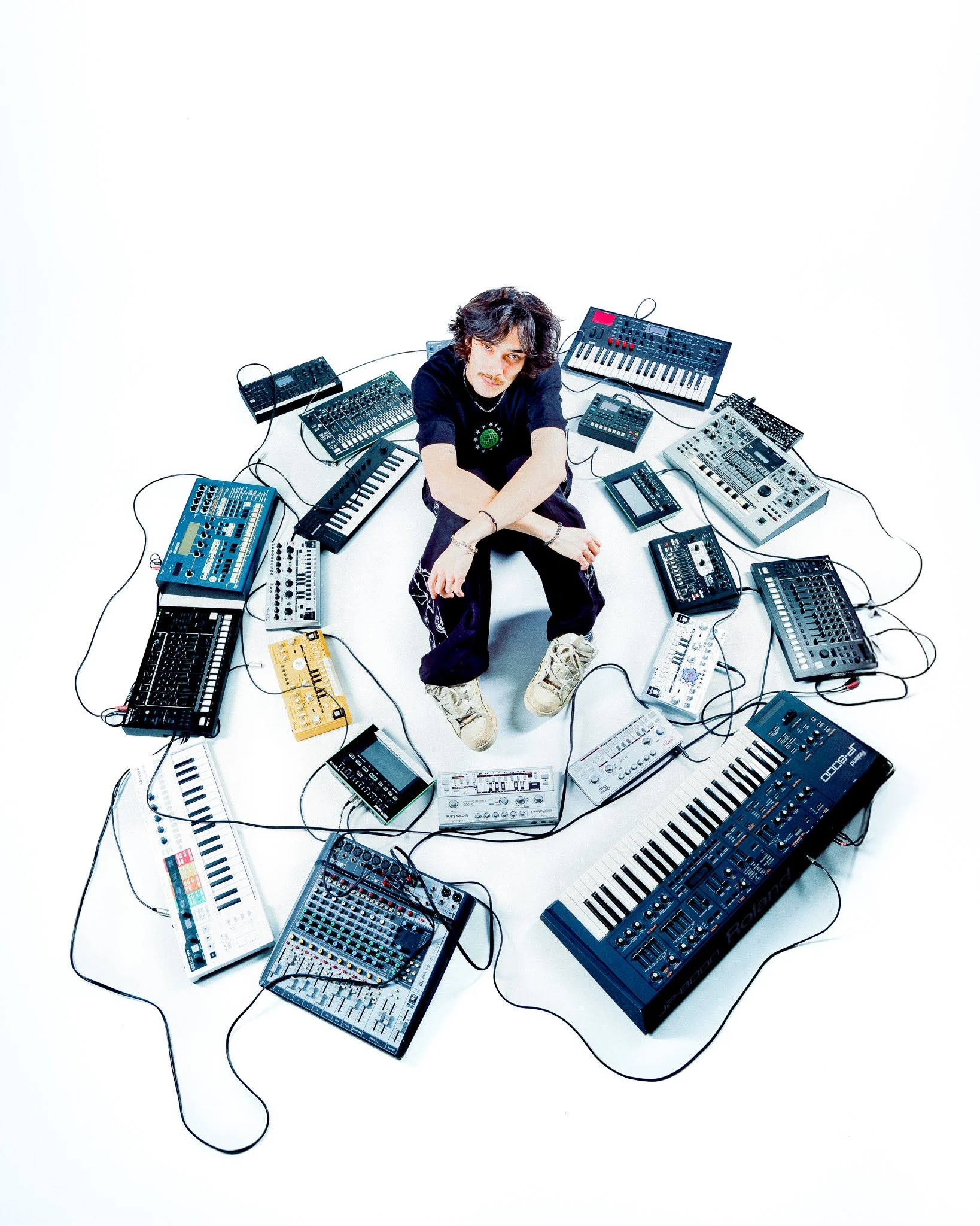 Jeune homme assis au centre entouré de divers équipements de musique électronique, notamment des claviers, des synthétiseurs, des tablettes et des contrôleurs, tous connectés par des câbles.