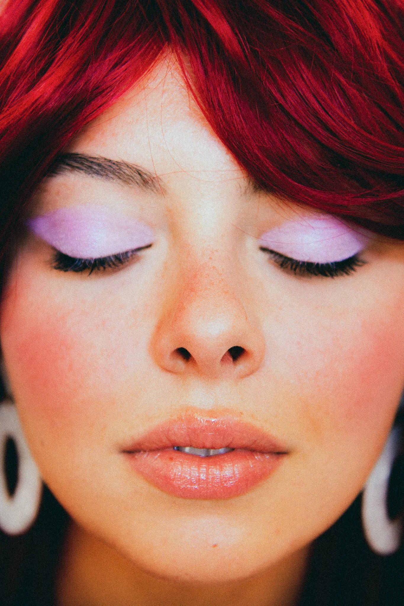 Femme avec maquillage pastel violet sur les paupières, cheveux rouges, boucles d'oreilles blanches, visage fermé, yeux fermés.