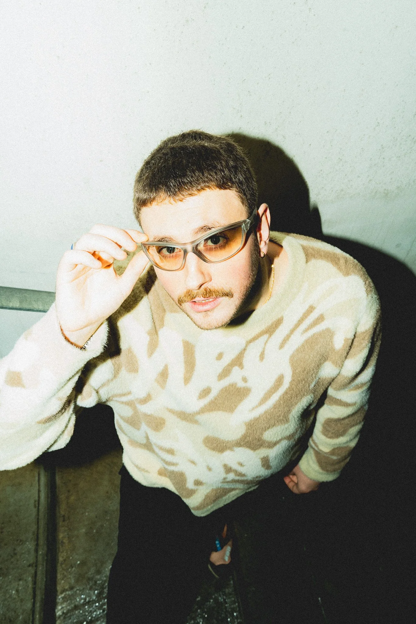 Jeune homme portant des lunettes de soleil et un pull beige à motifs, regardant vers l'appareil, dans un environnement intérieur.