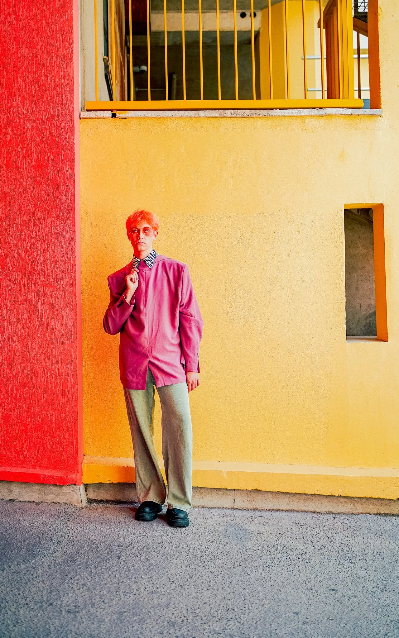 Une personne debout devant un mur jaune avec un panneau rouge à gauche, portant un blazer rose, une chemise à motifs et un pantalon beige, dans un environnement coloré et moderne.