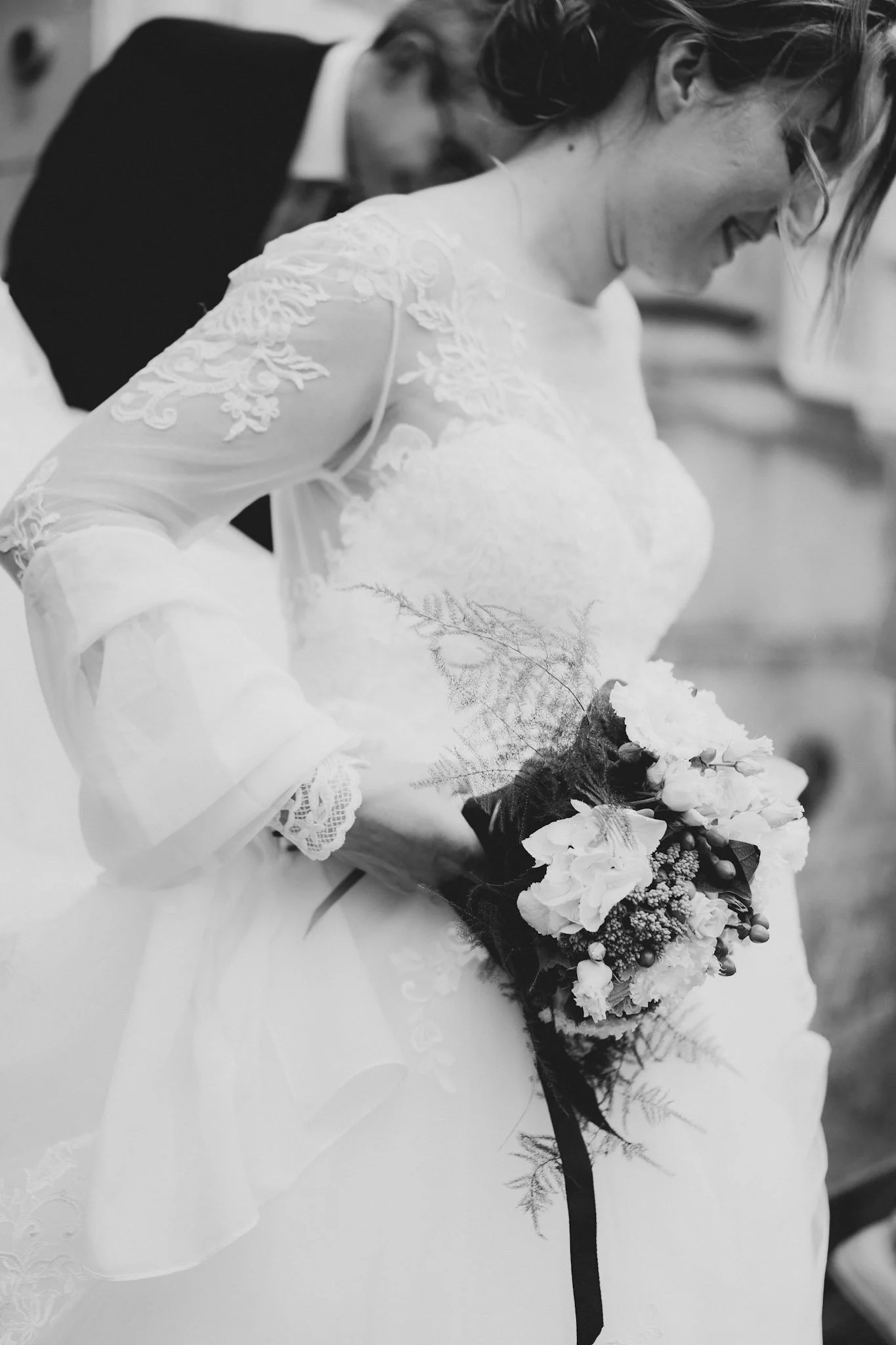 Une mariée en robe blanche avec des broderies en dentelle, tenant un bouquet de fleurs, souriant, lors d'une cérémonie. En arrière-plan, un homme en costume sombre. La photo est en noir et blanc.