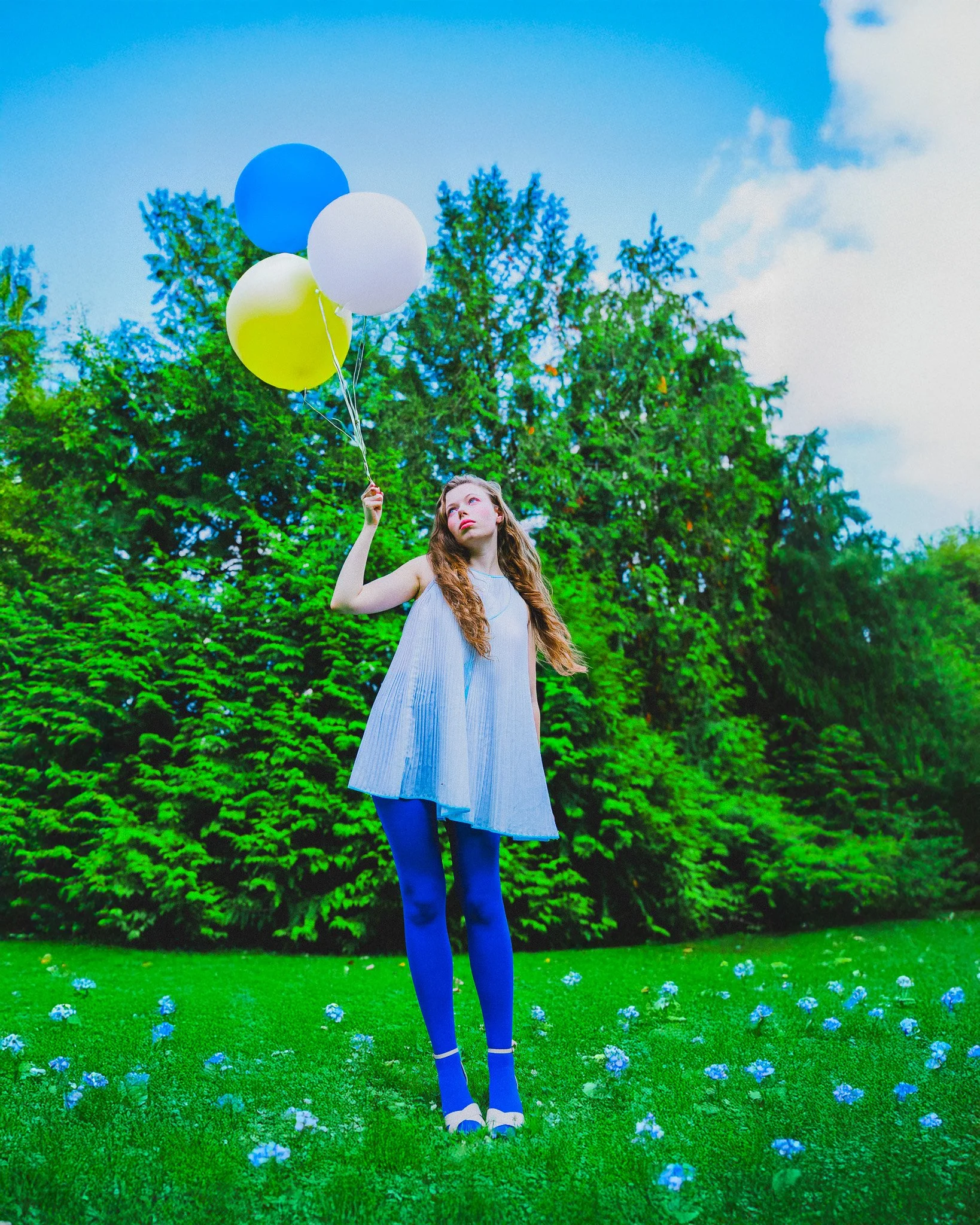 Jeune femme avec des collants bleus et une robe blanche, tenant des ballons colorés dans un parc vert.