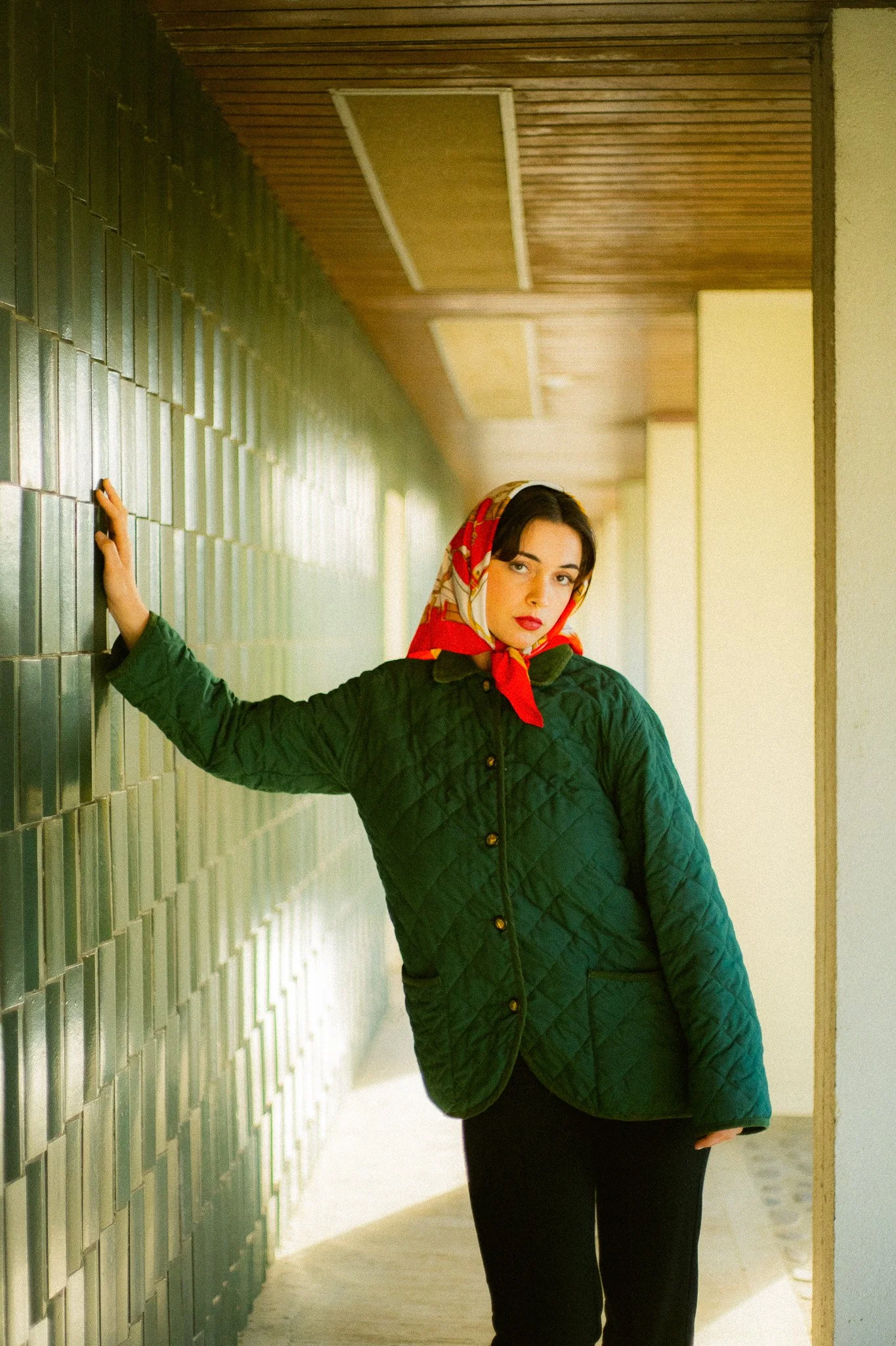 Femme portant une veste verte et un foulard rouge, posant dans un couloir intérieur avec murs en carreaux verts et plafond en bois.