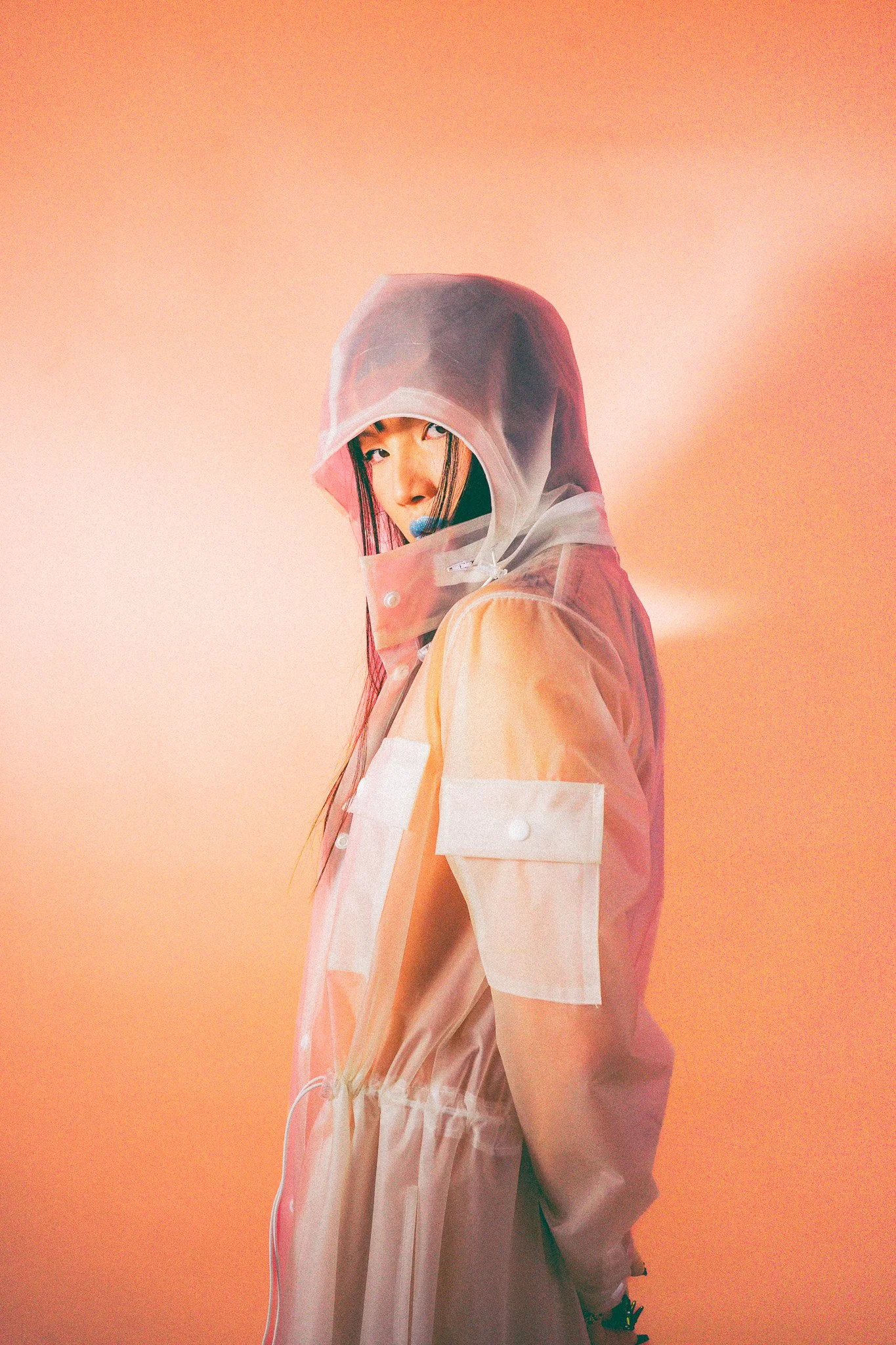 Femme portant un manteau transparent avec capuche, regardant par-dessus son épaule, fond orange
