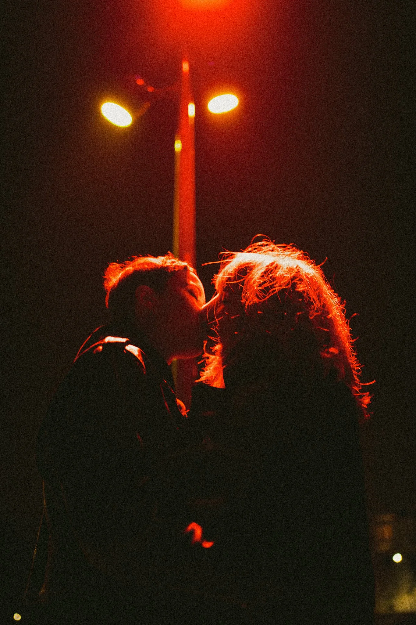 Une scène de nuit avec deux personnes en gros plan, une d'elles kissant l'autre, sous un lampadaire rougeâtre.