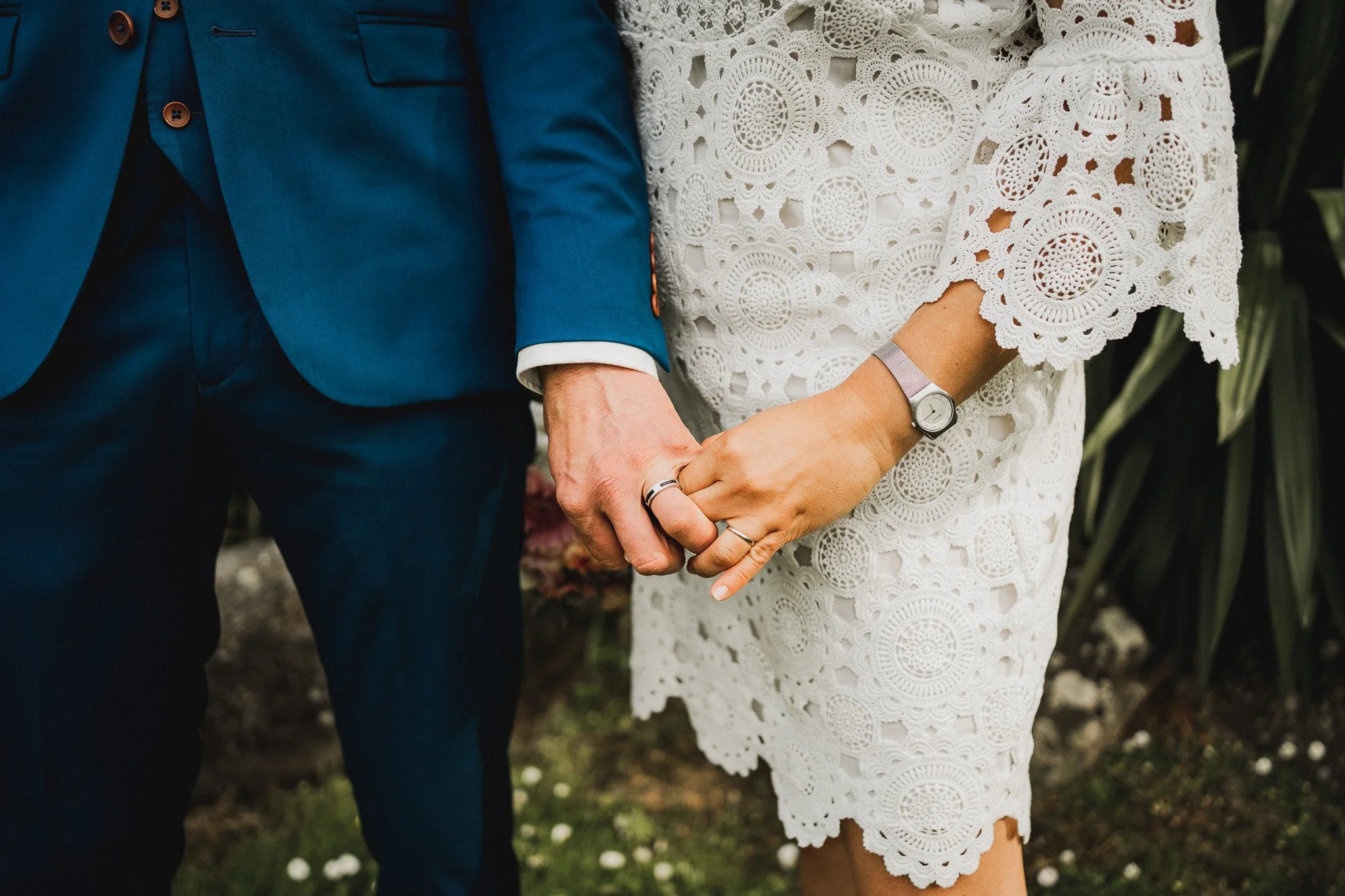 Un couple se tenant la main lors d'une cérémonie, lui en costume bleu, elle en robe blanche en crochet, portant une montre et des alliances.