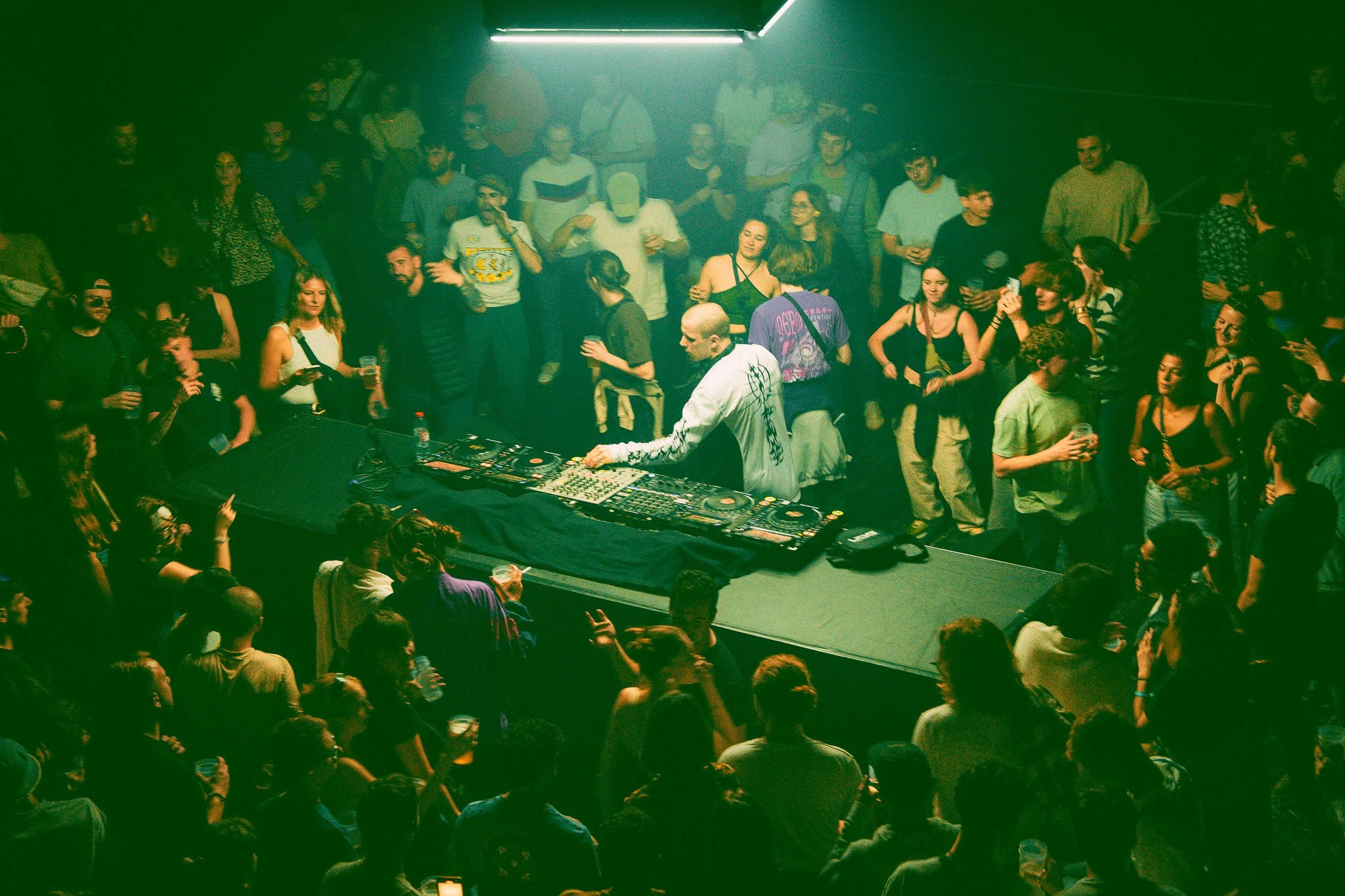 Un DJ joue de la musique dans une boîte de nuit animée avec une grande foule de personnes qui dansent et boivent des boissons, éclairée par des lumières vertes.