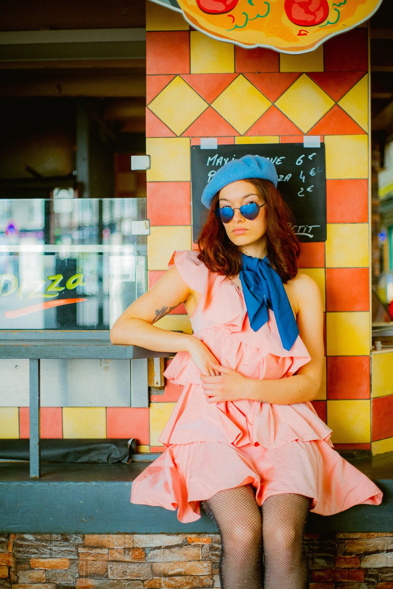 Une jeune femme assise dans une pizzeria, portant une robe rose, un béret bleu, des lunettes de soleil rondes, et un nœud bleu au cou, avec un mur de briques jaunes et oranges en arrière-plan.
