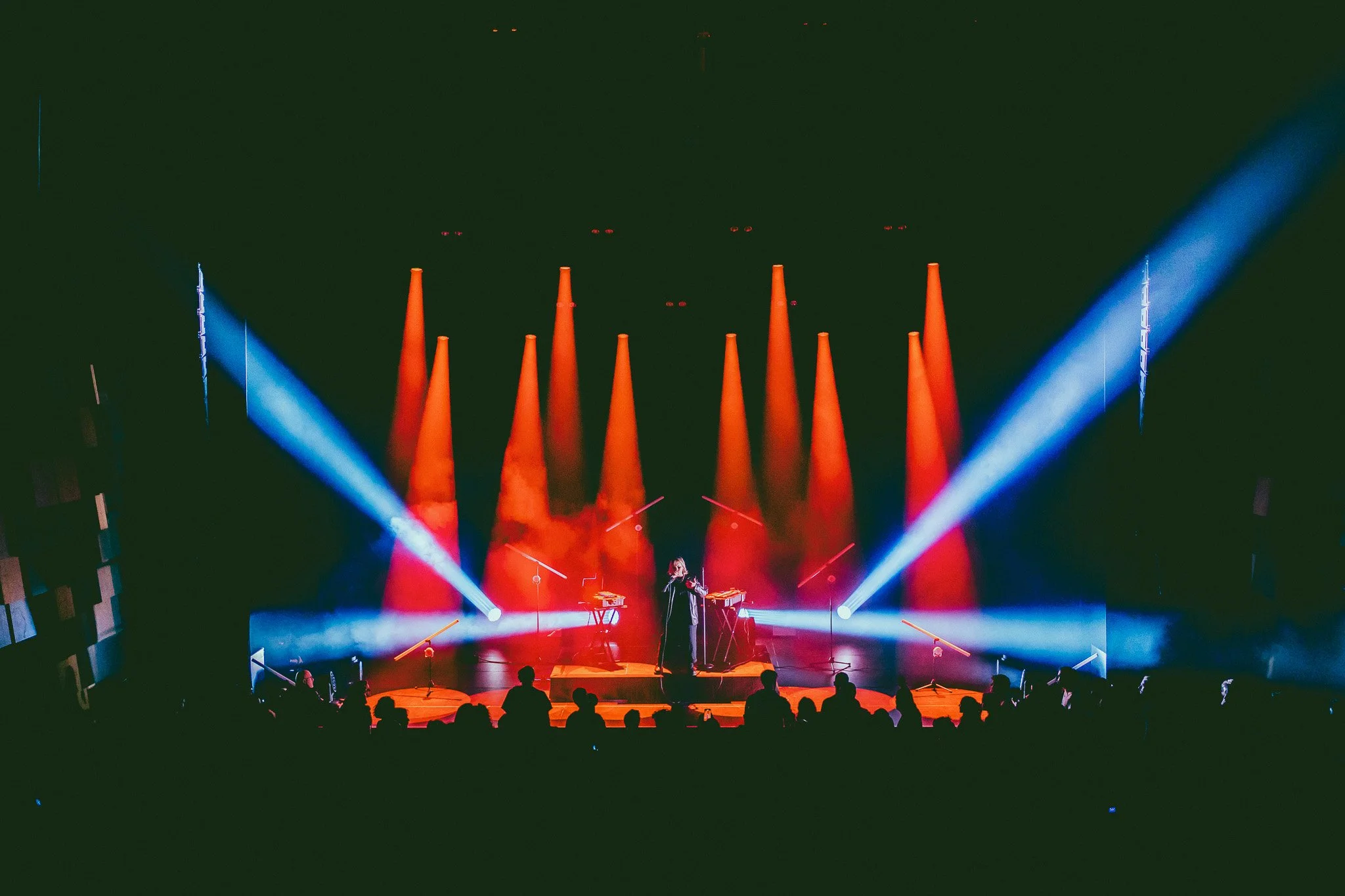 Une scène de concert avec un artiste sur scène, entouré de lumières bleues et orange, et un public en silhouette devant.
