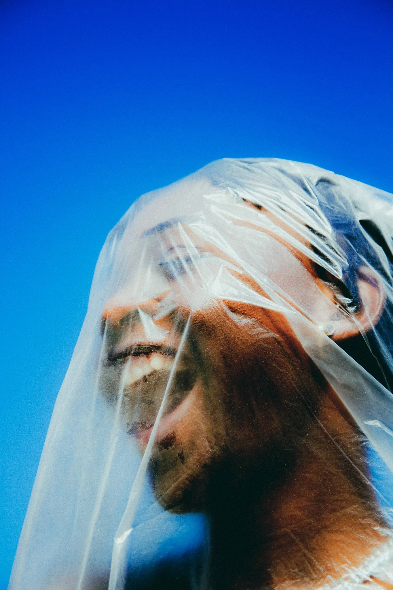Un portrait d'une femme avec un voile en plastique transparent sur la tête, souriant légèrement, contre un ciel bleu clair.