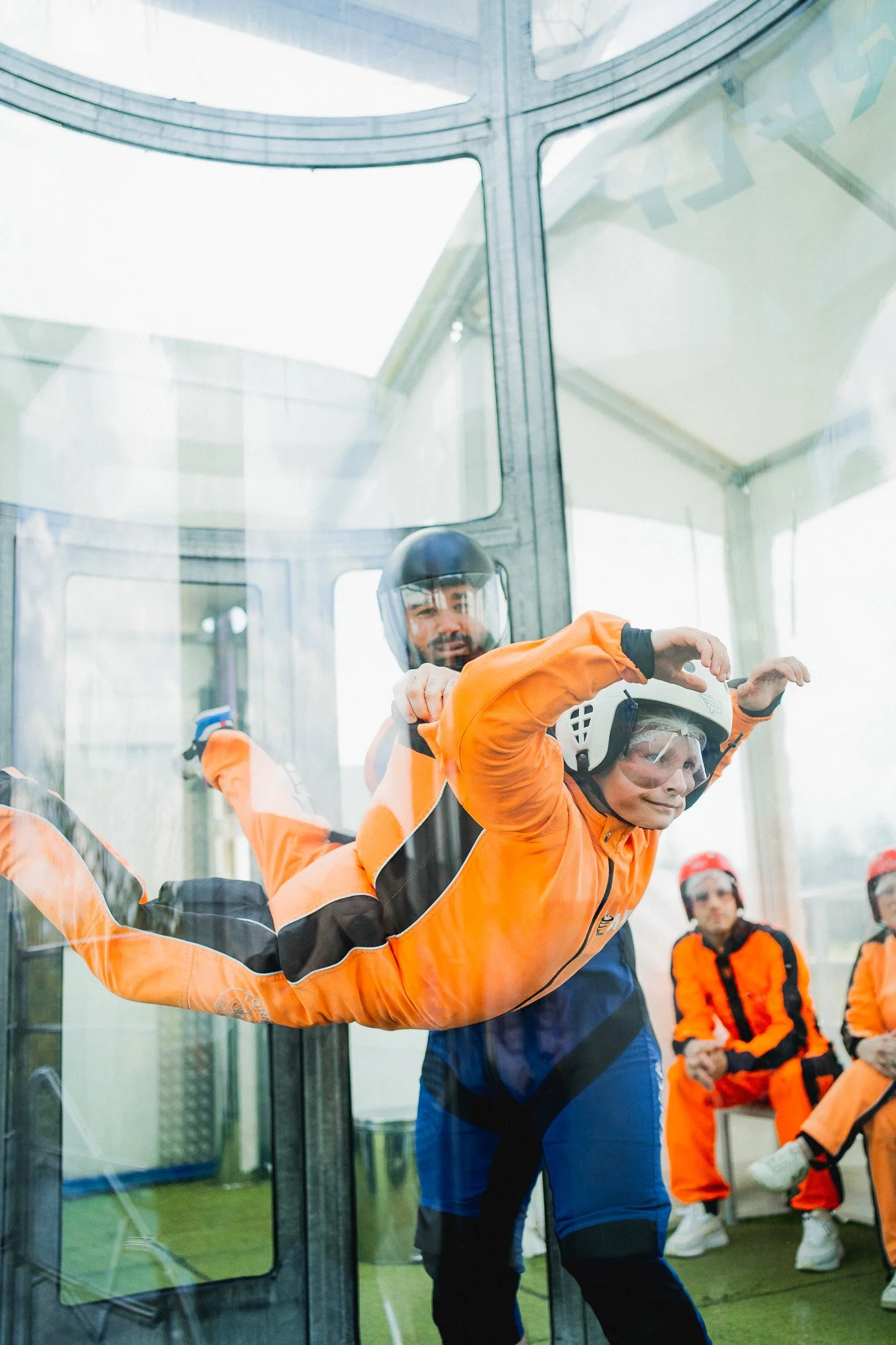 Un instructeur aide un participant à faire un saut en parachute à l'intérieur d'une corde à trampoline. Tous deux portent des combinaisons orange et des casques de protection, avec d'autres personnes déguisées en sauteurs en parachute en arrière-plan.