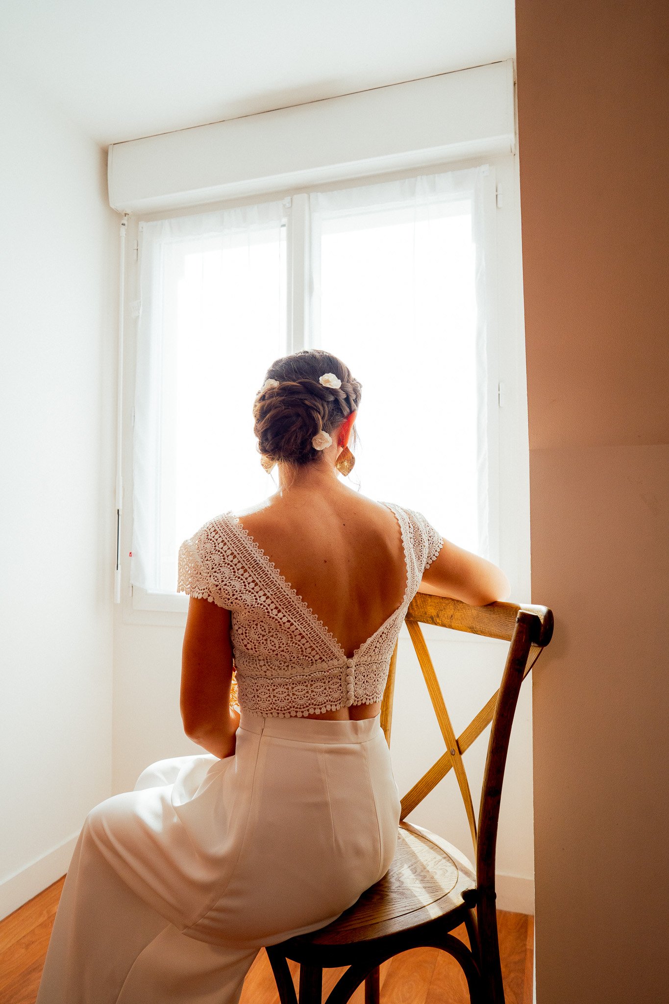 Une femme assise sur une chaise en bois, vue de dos, regardant une fenêtre lumineuse, portant une blouse en dentelle blanche avec le dos dénudé et une jupe beige, avec des fleurs dans ses cheveux.