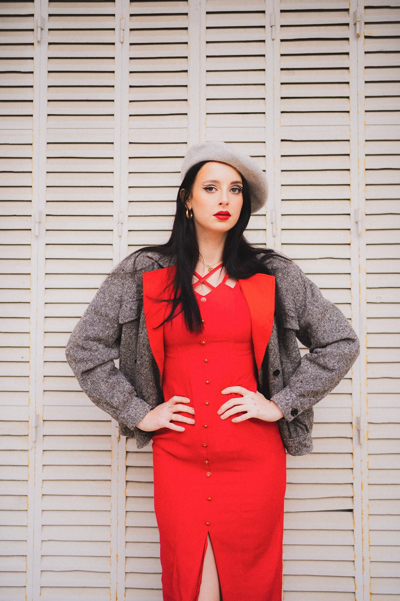 Femme portant une robe rouge et un manteau à carreaux gris, avec un chapeau gris, posant devant un panneau blanc à lattes.