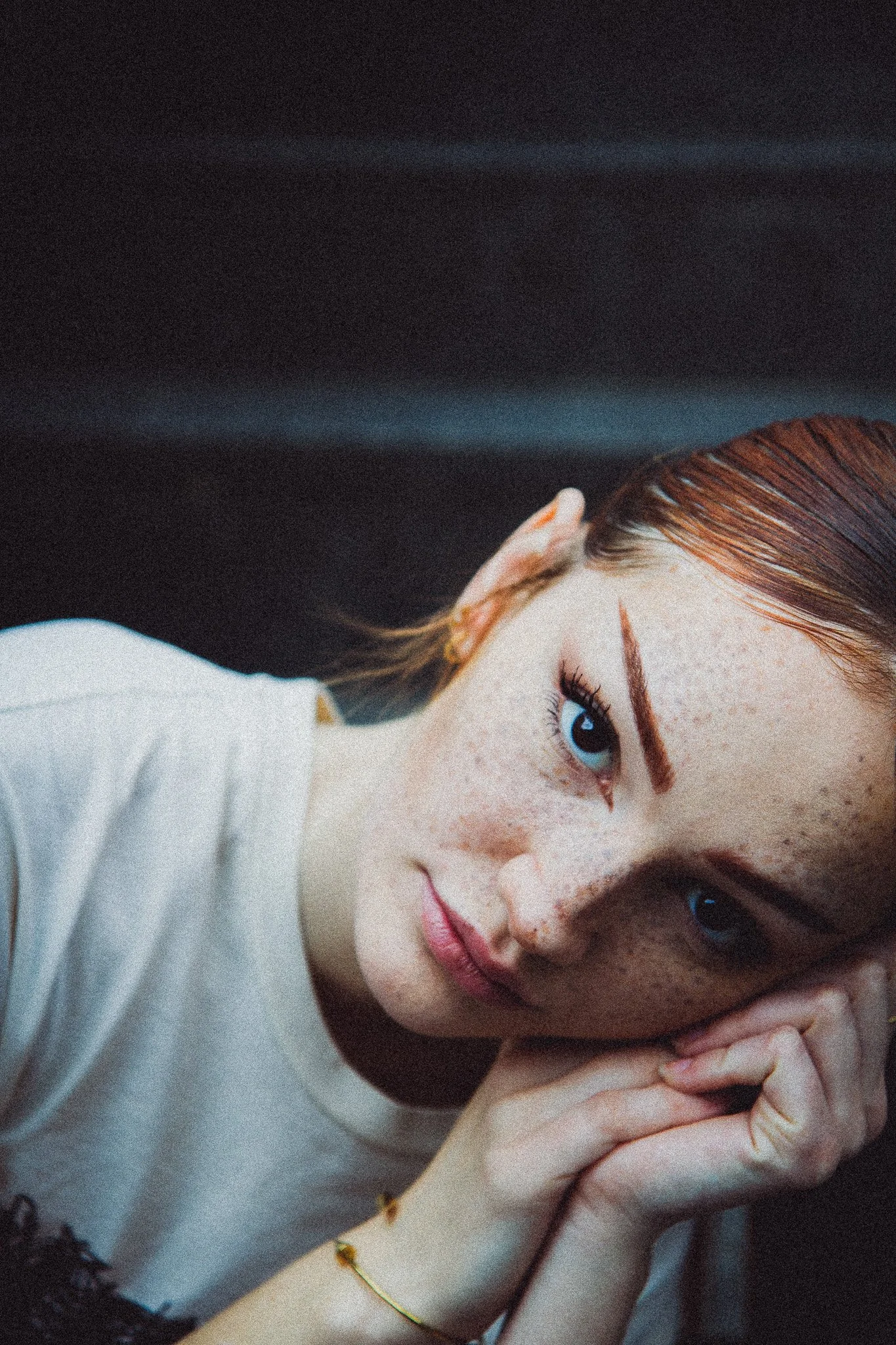 Jeune femme aux cheveux roux, yeux bleus, t-shirt blanc, en train de se reposer sur une main, fond sombre.