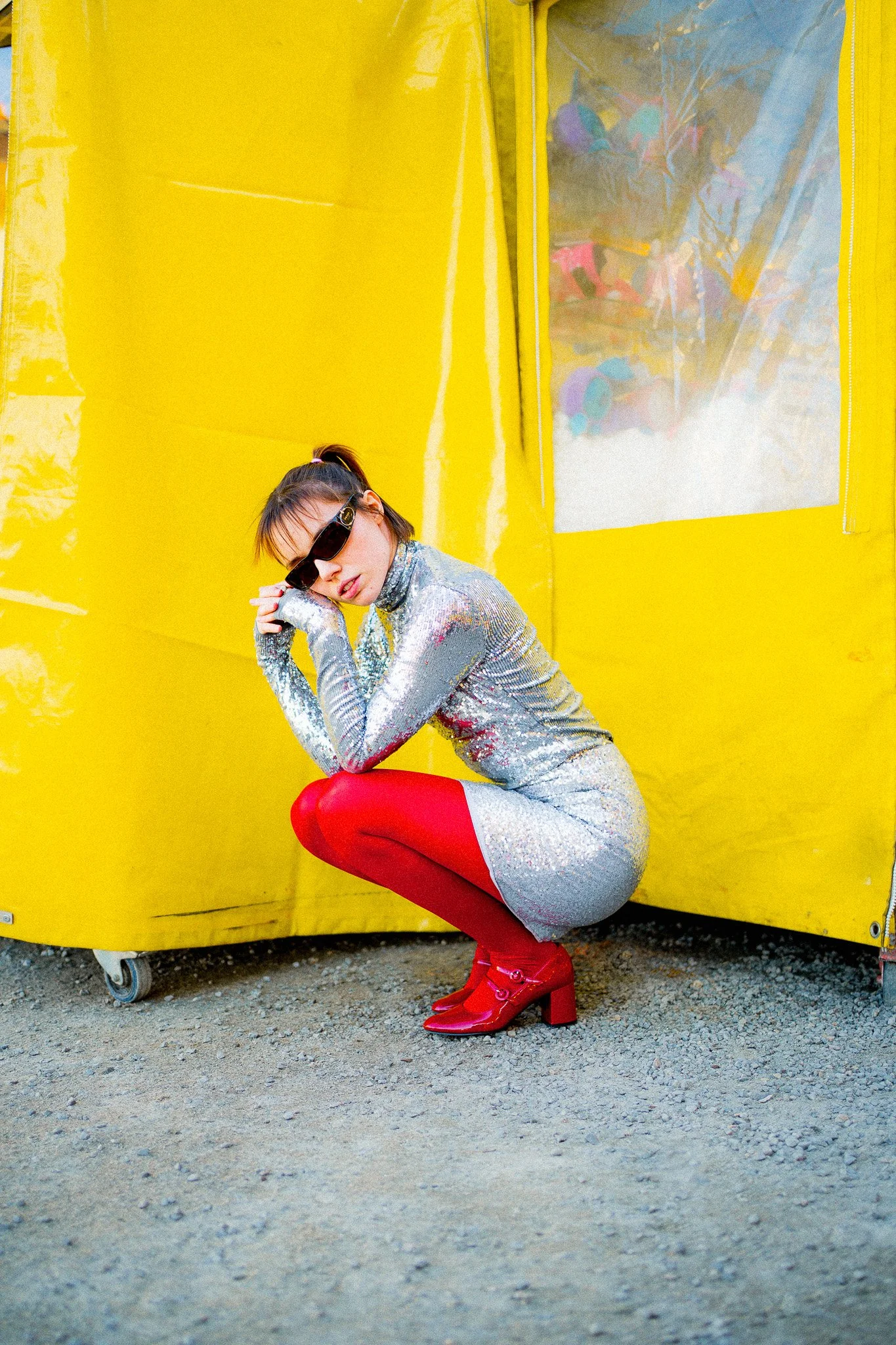 Jeune femme fashion portant une robe argentée en sequins, des collants rouges et des bottes rouges, posant devant une structure jaune avec une fenêtre en plastique.