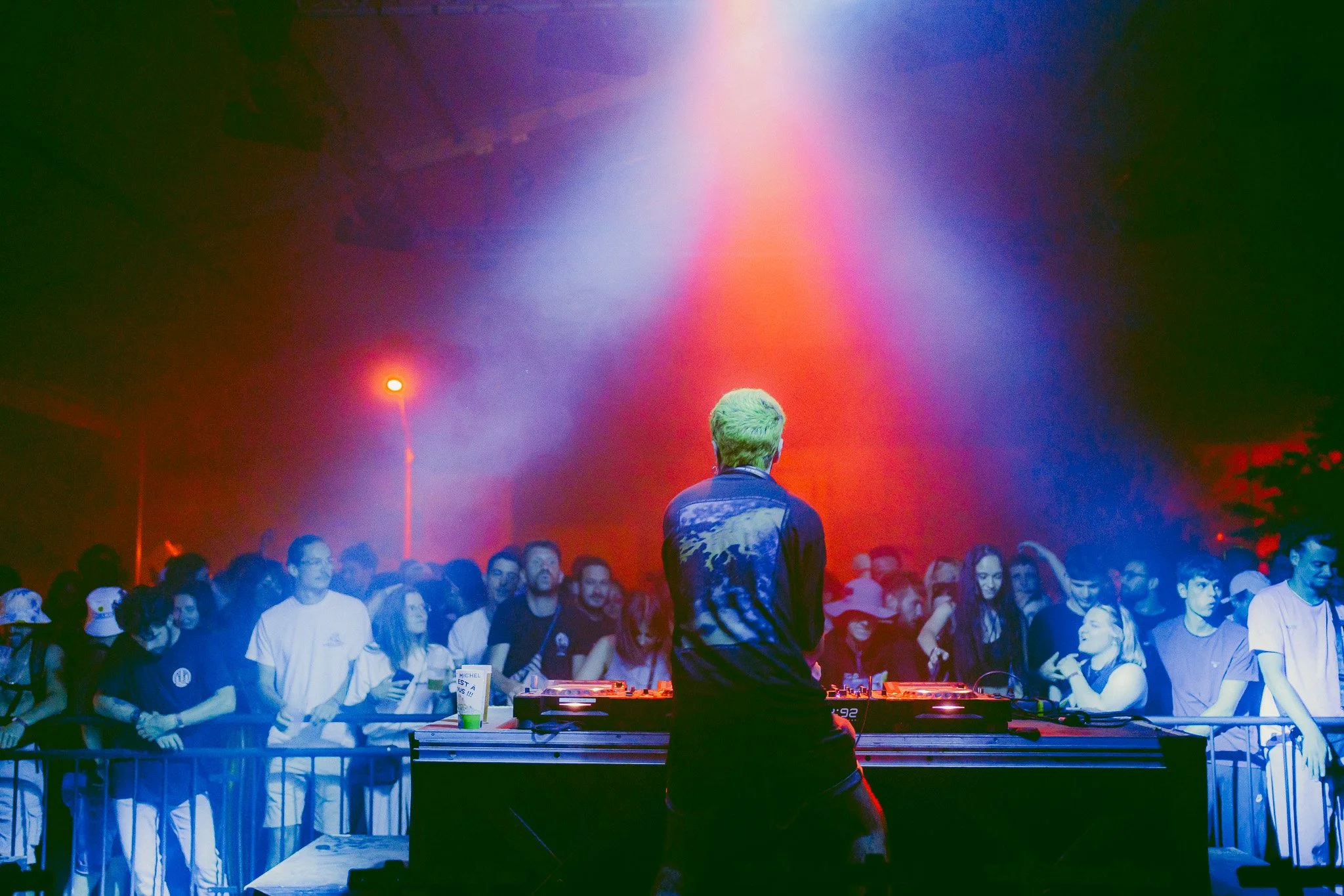 Un DJ joue de la musique devant un groupe de spectateurs dans une fête en plein air, avec des lumières colorées et un ciel nocturne.